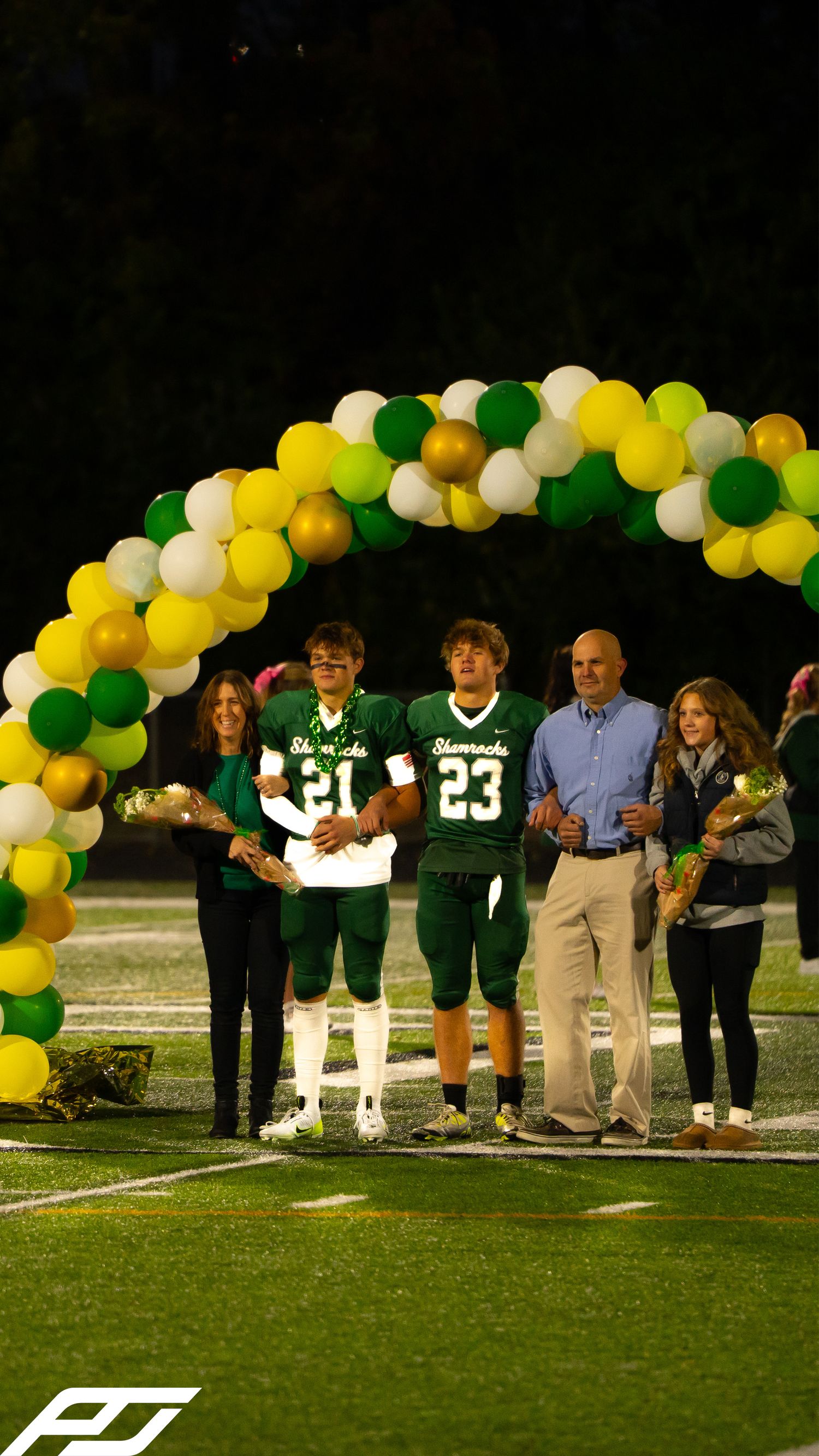Shamrocks football players #21 and #23 stand with families under balloon arch during senior night celebration.