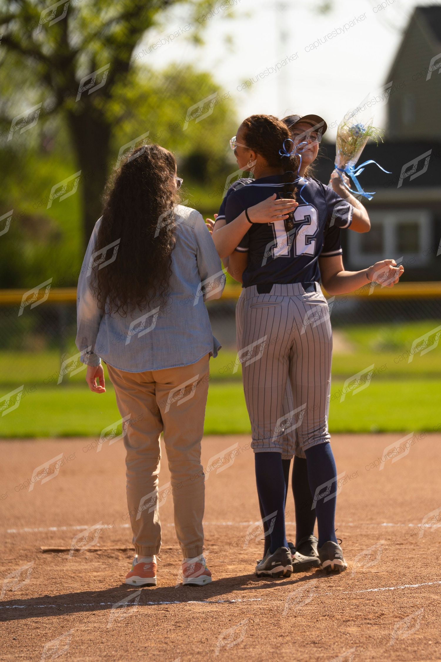 Softball player wearing number 12 receives warm hug from teammate during senior day celebration on field.