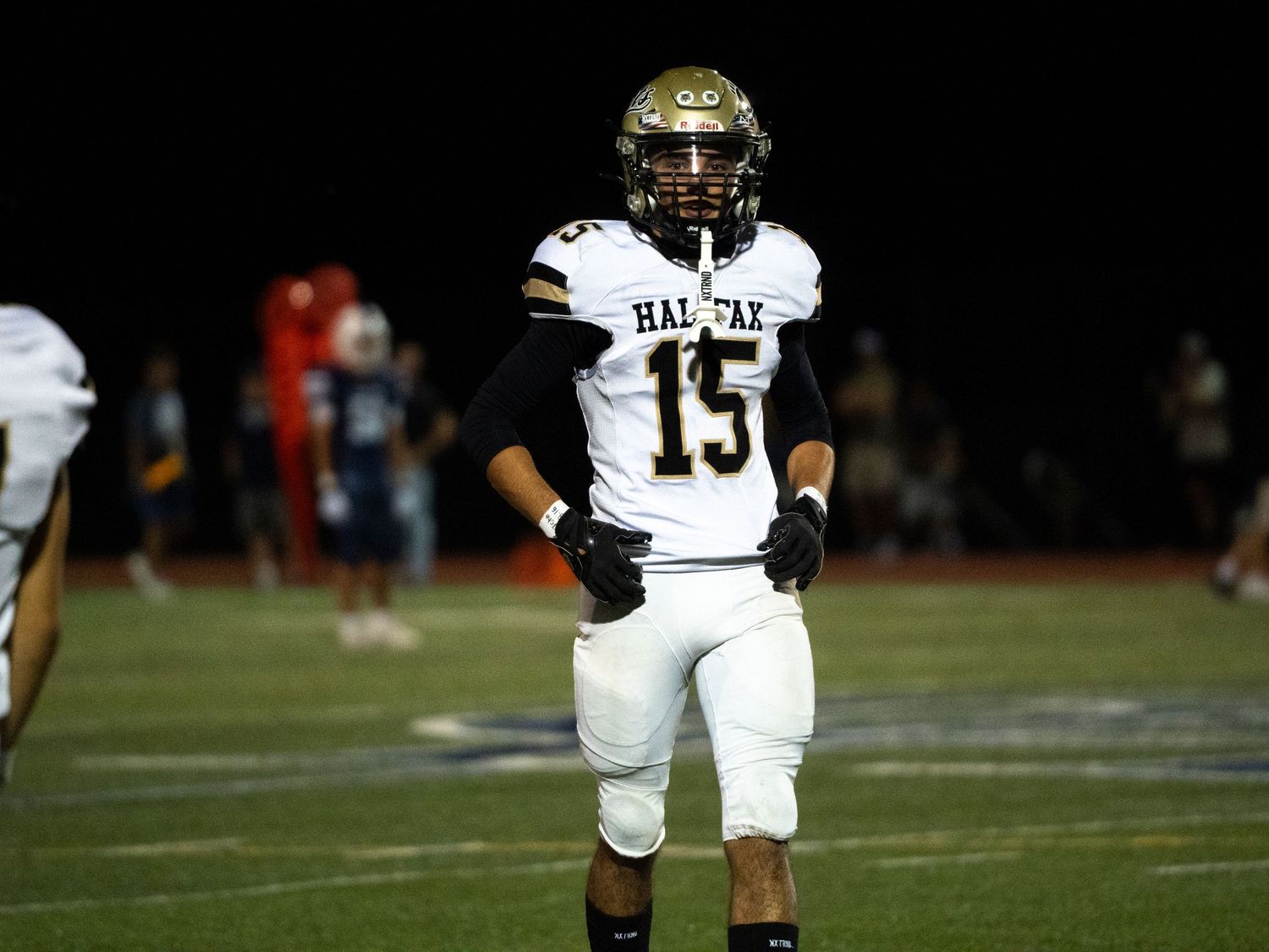 A Halifax high school football player wearing number 15 jogs on the field during a night game in gold and black uniform.
