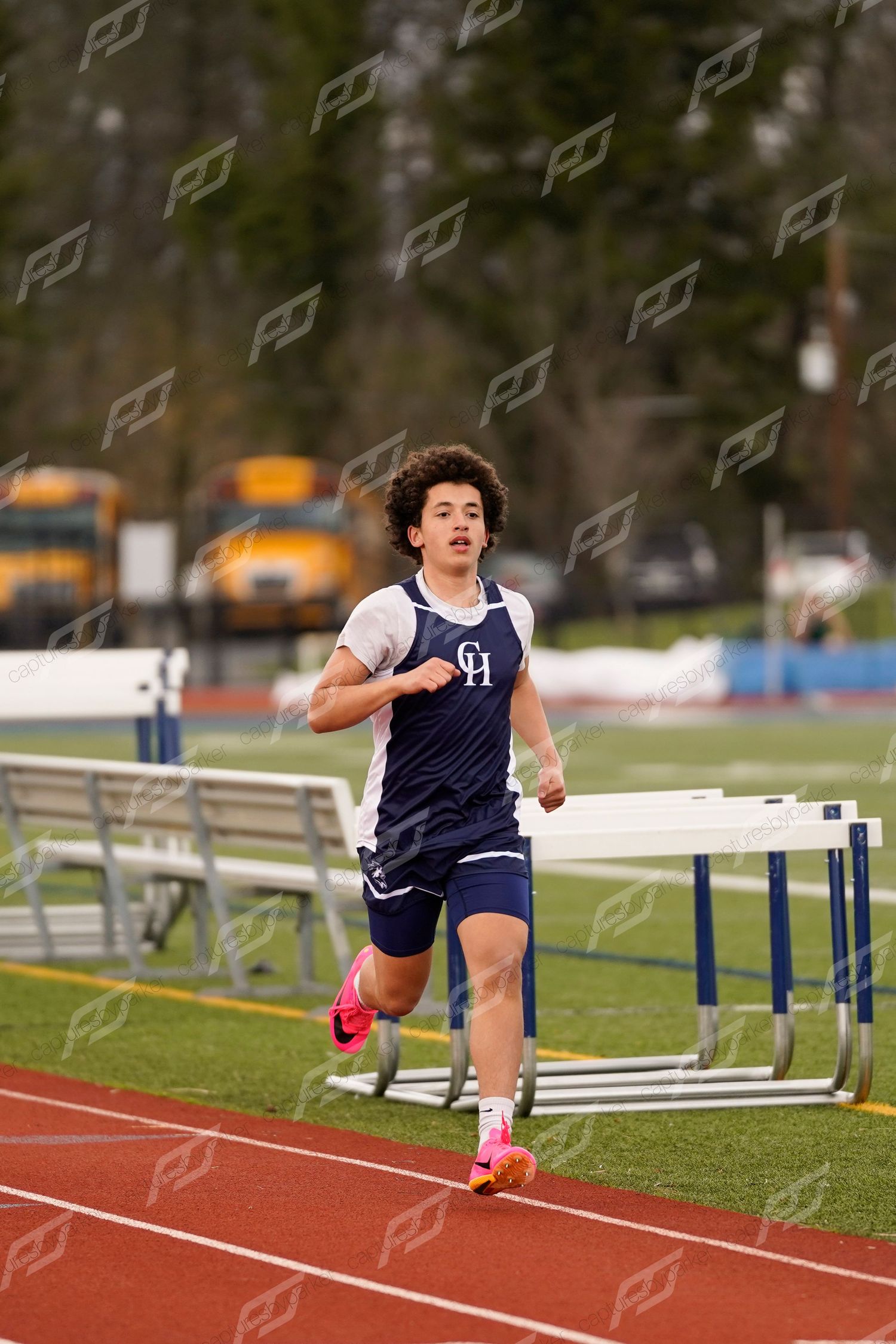 Curly-haired male athlete in navy uniform running on a red track past white hurdles.