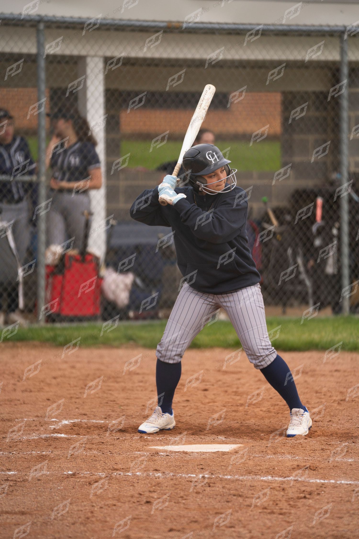 Female softball player in batting stance wearing navy uniform awaits pitch at home plate.