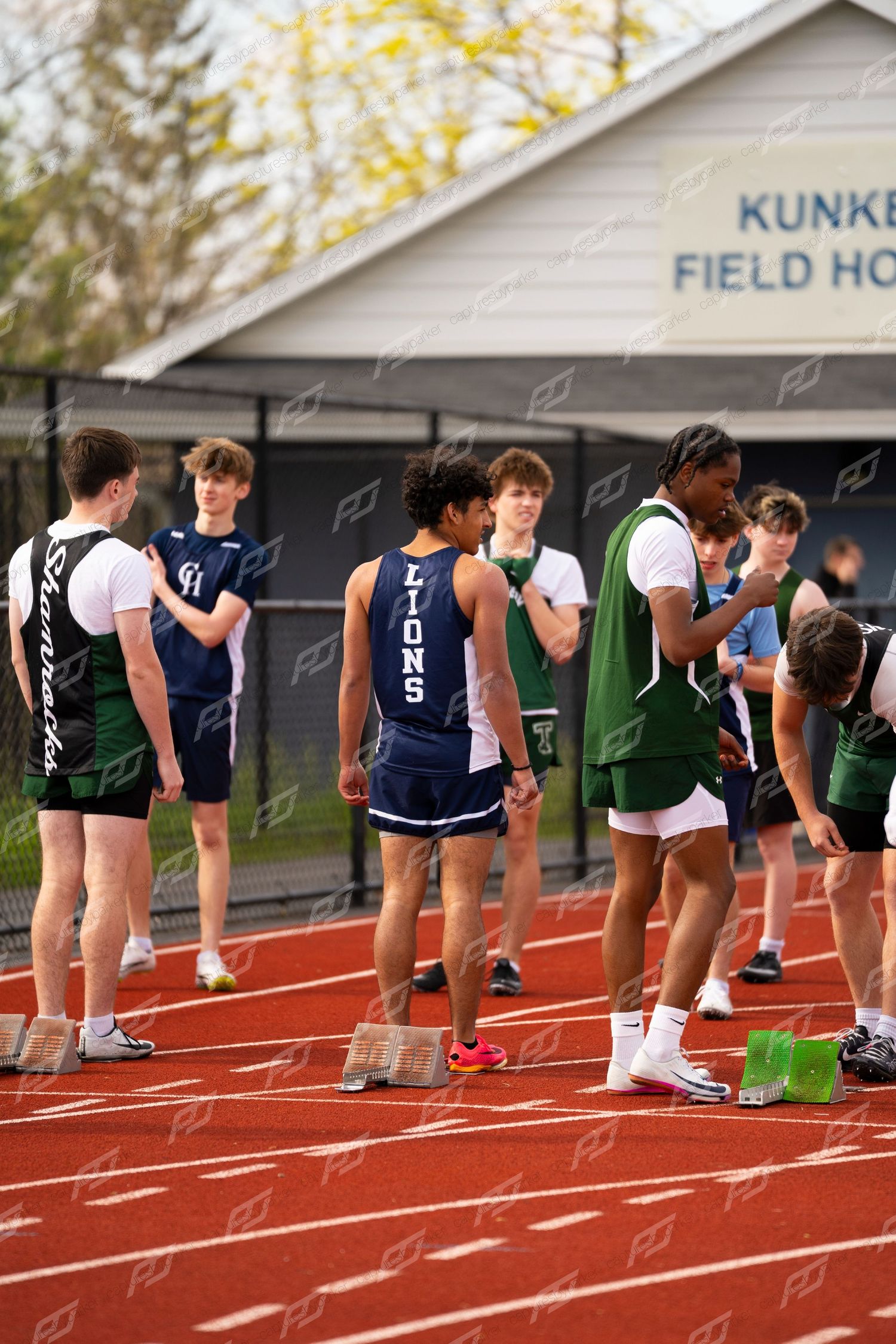 Male high school track athletes in Lions and green uniforms gather on red track at Kunkel Field House.
