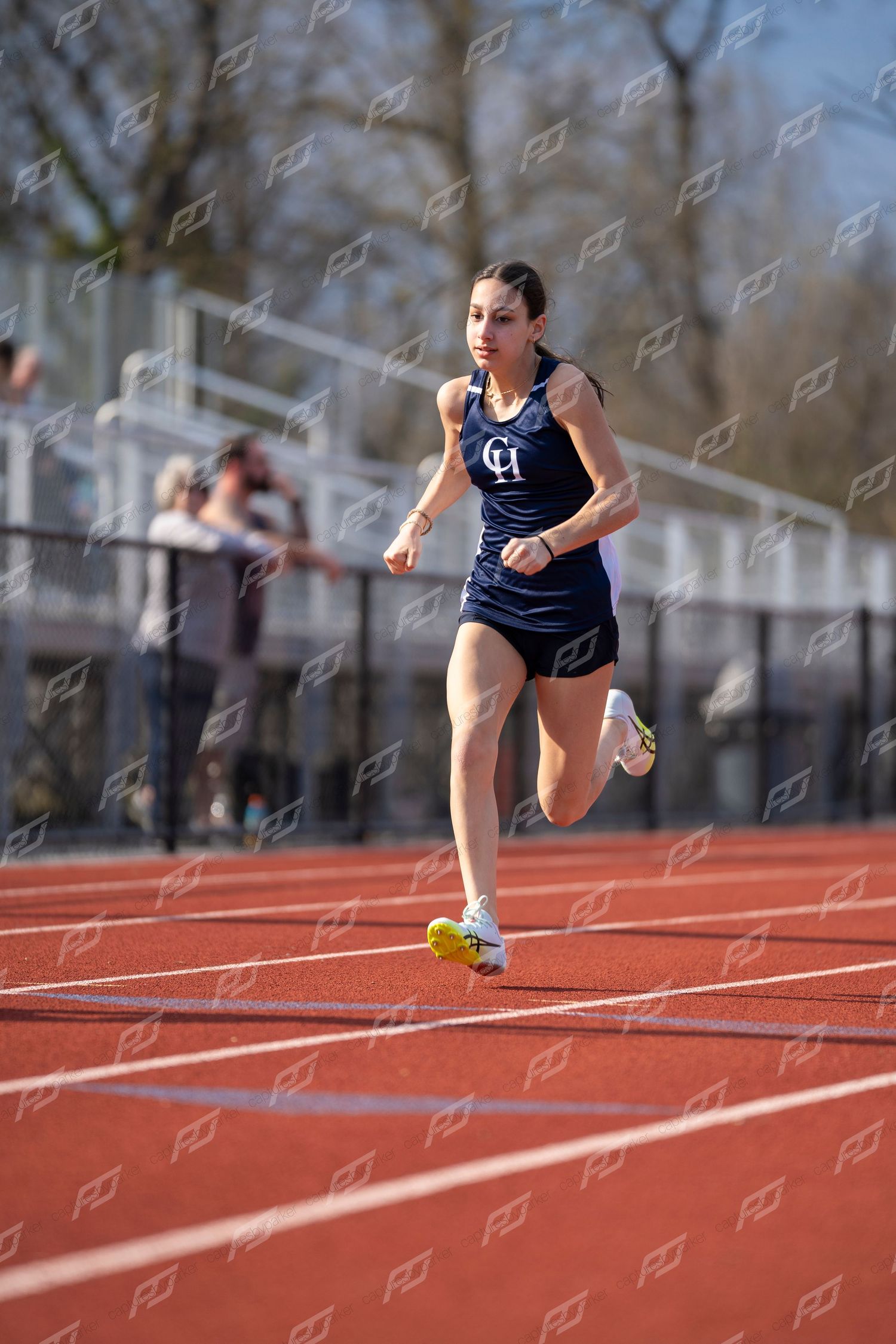 Female high school sprinter in navy CH uniform runs on red outdoor track.