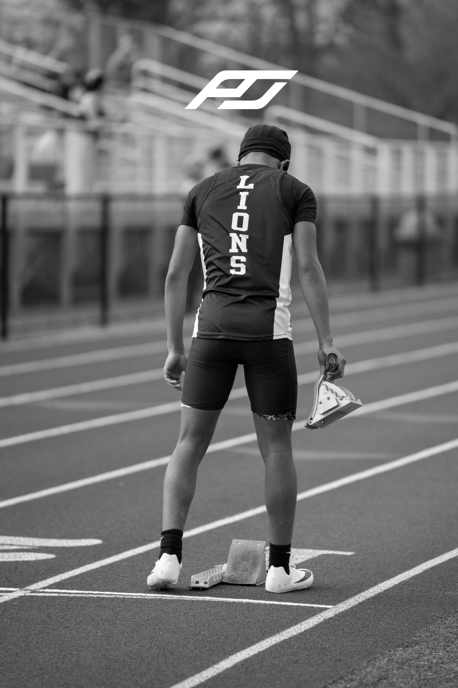 Lions sprinter stands at starting block holding spike shoes on track in black and white.