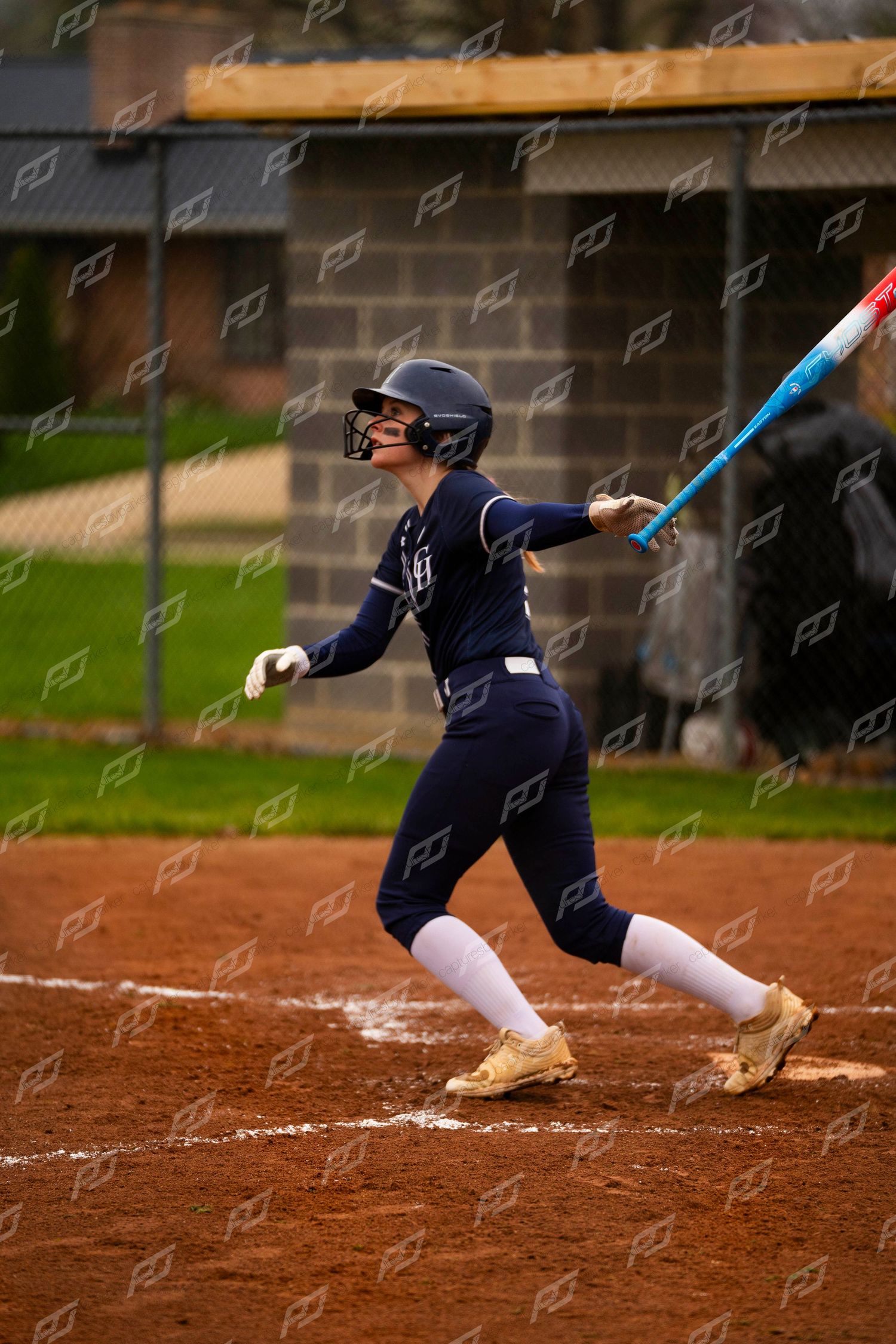 Female softball player in navy uniform completes a full follow-through swing with a colorful bat on dirt field.