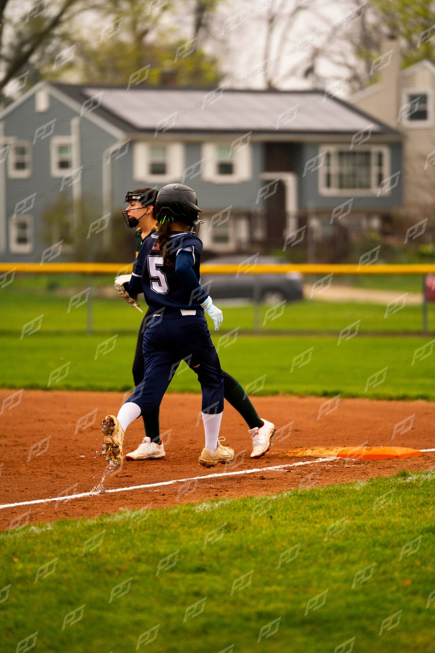 Two softball players in navy uniforms run bases on a red dirt field with a house visible in the background.