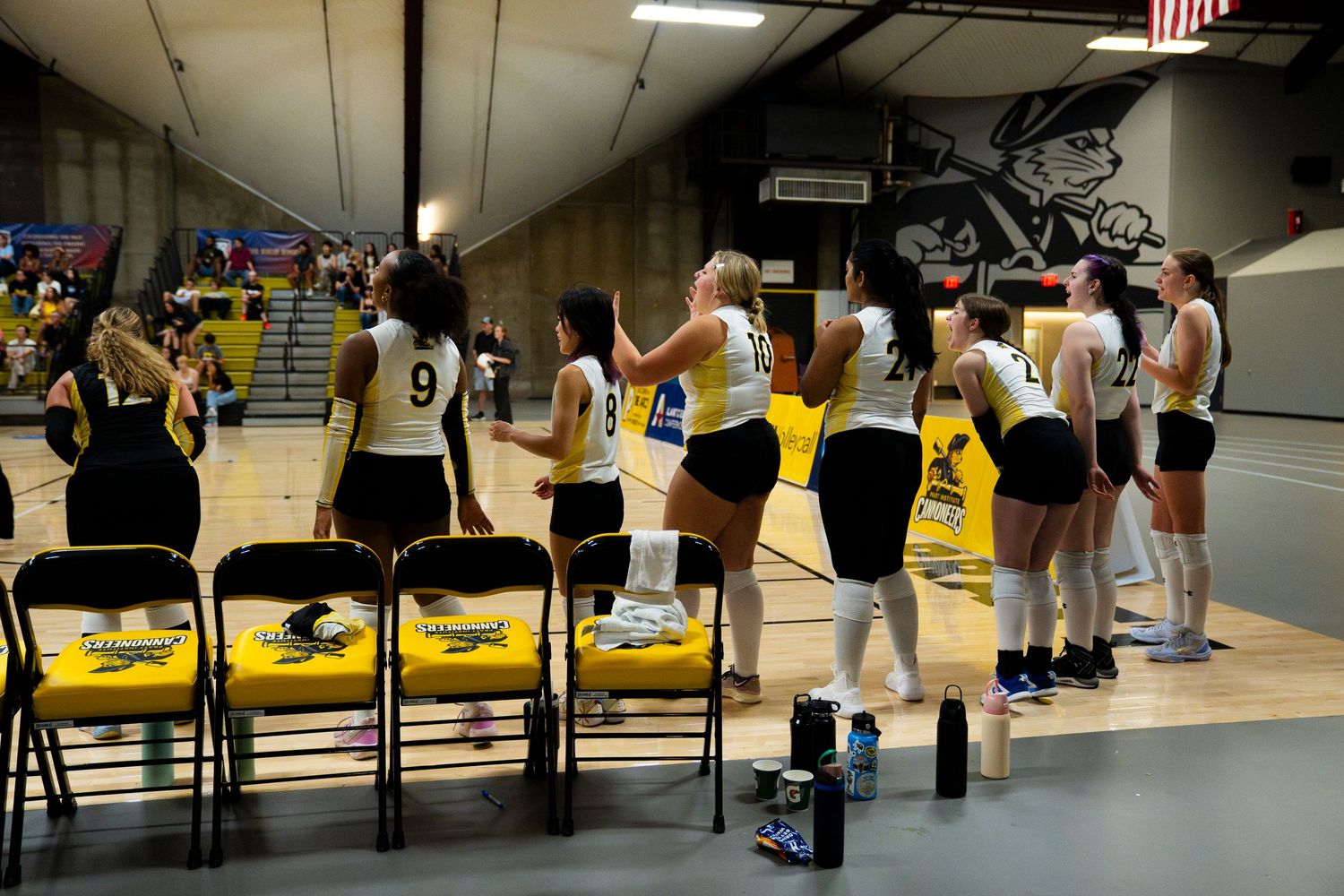 Women's volleyball team in black and yellow uniforms standing courtside during a game in an indoor gymnasium.