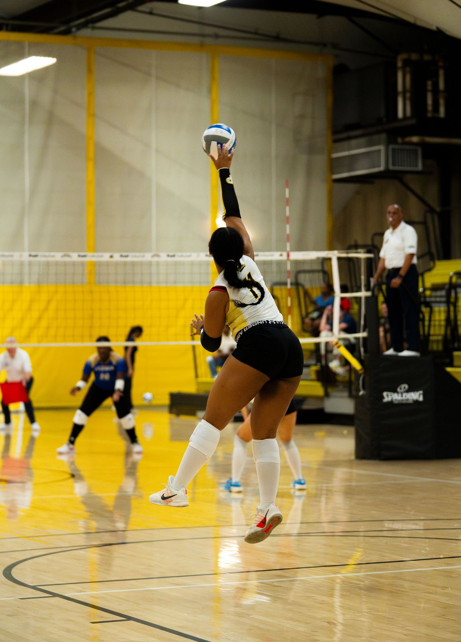 Female volleyball player jumping to spike the ball during an indoor game on a hardwood court.