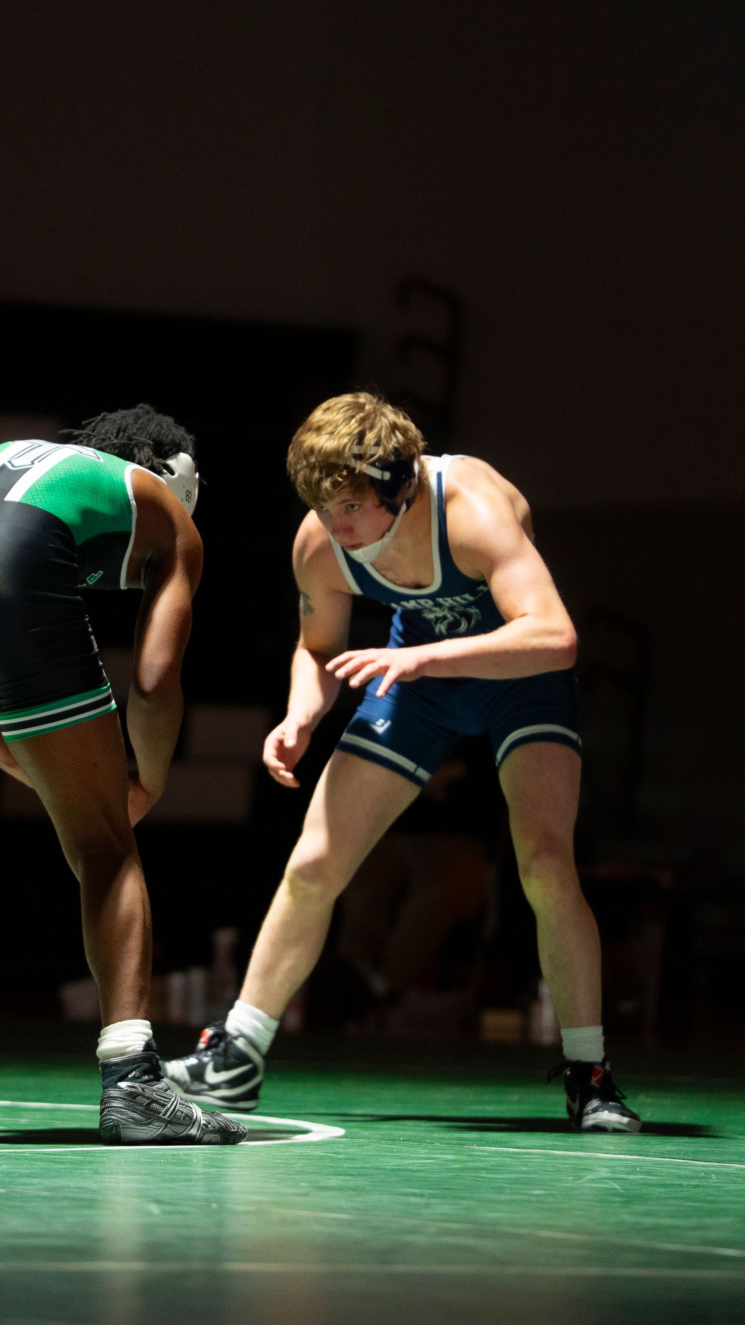 Two wrestlers in blue and green singlets face off in ready stance on mat.
