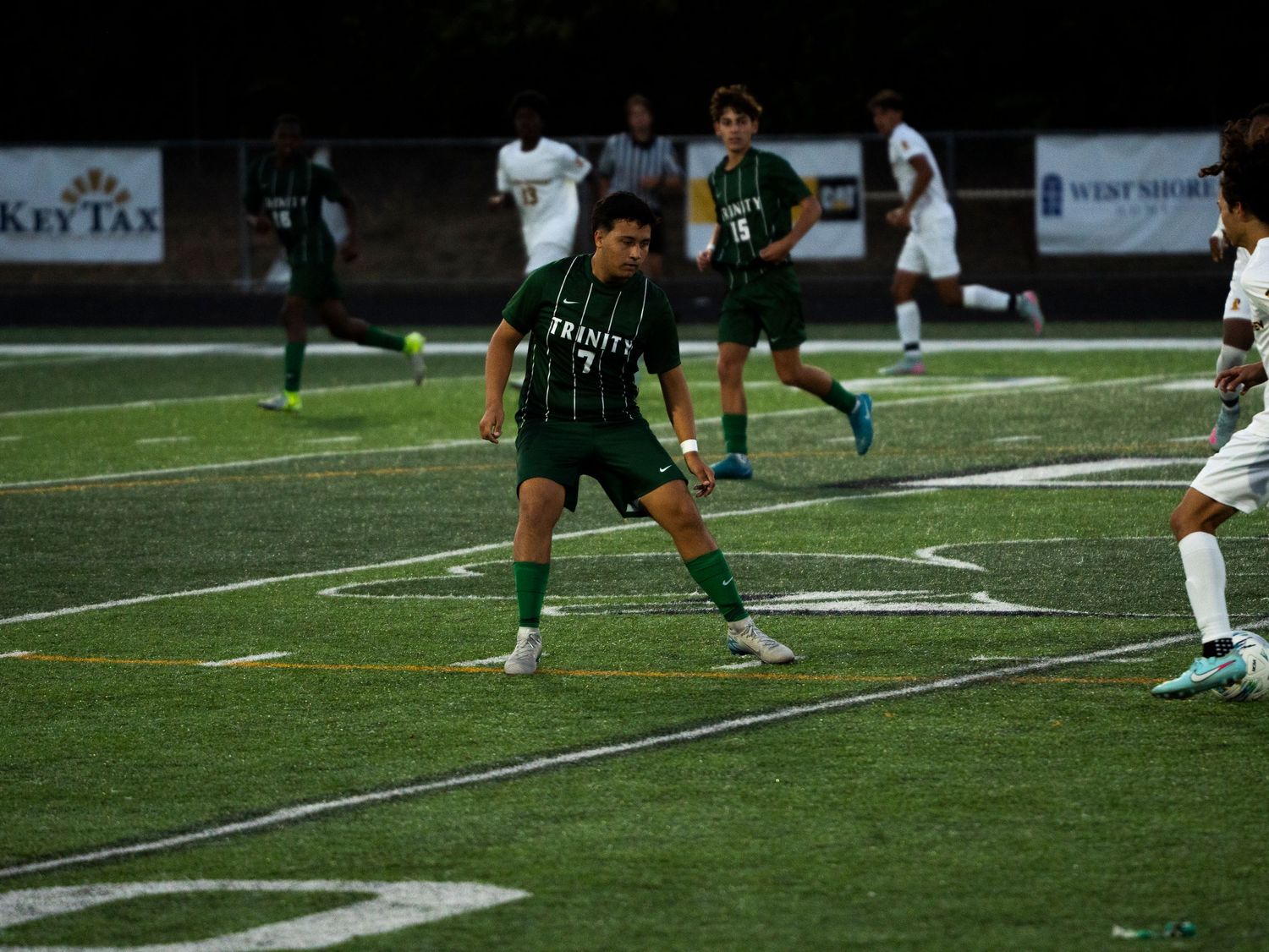 Trinity #7 soccer player dribbles ball during night game with teammates visible on lit turf field.