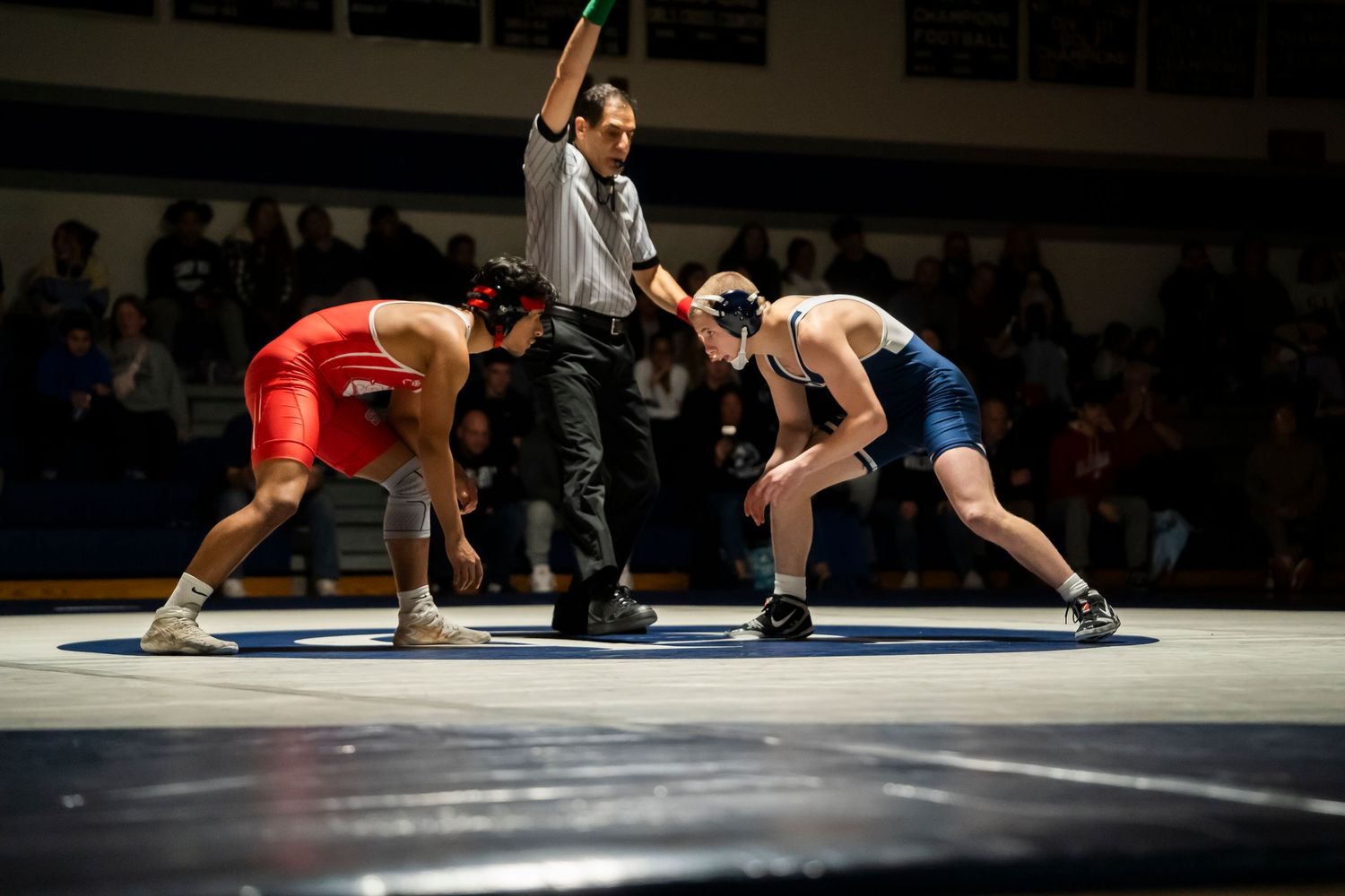 Referee raises hand as two wrestlers in red and blue singlets square off on the wrestling mat.
