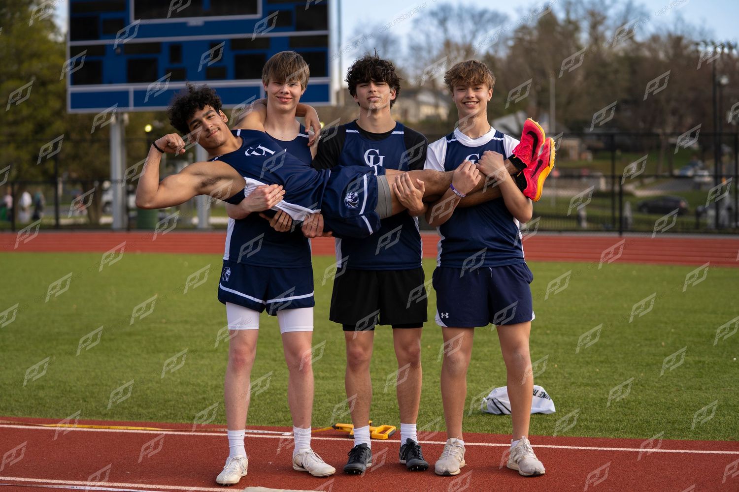 Four male high school track athletes in navy CH uniforms pose together on red track.
