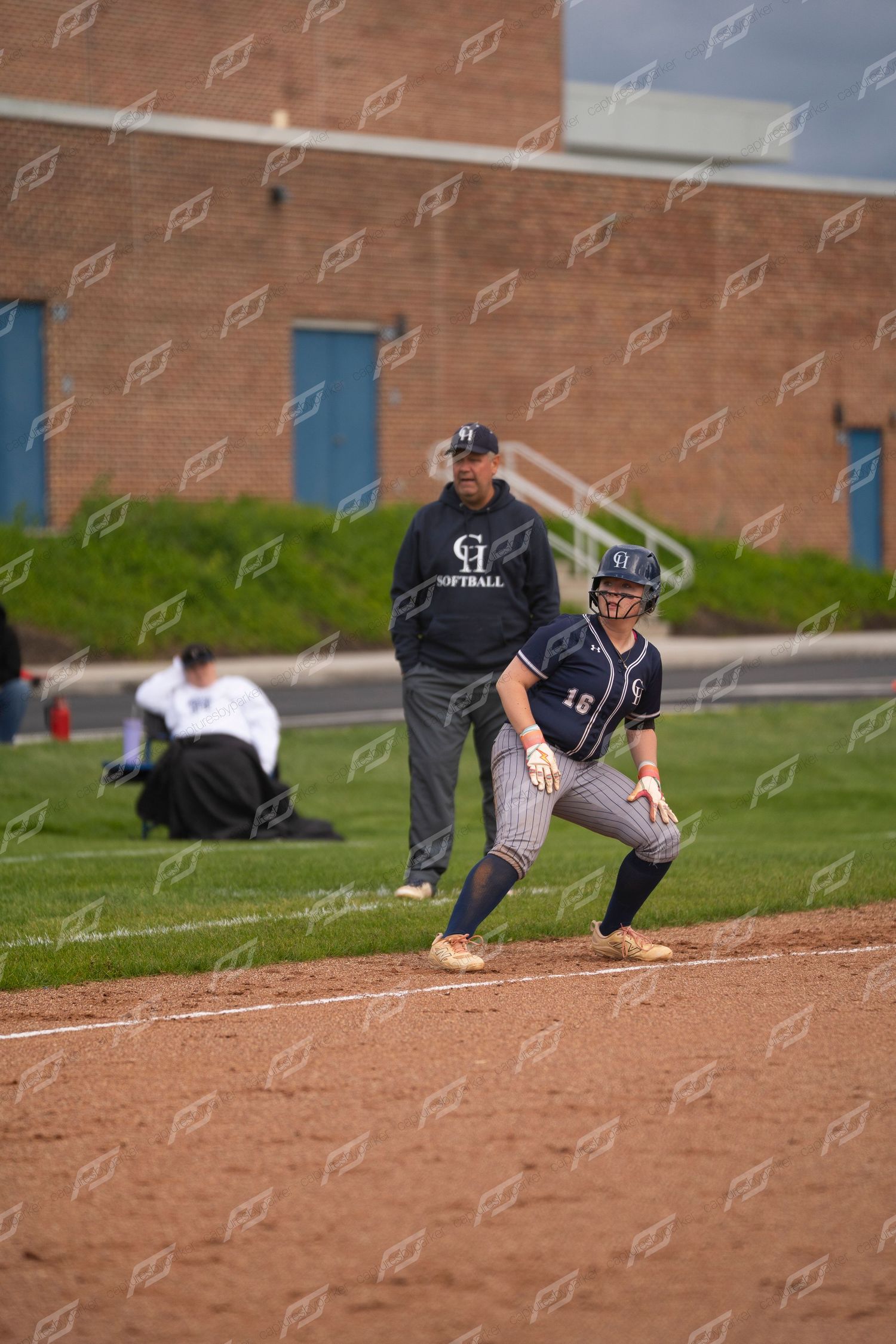 Softball player leads off first base as coach watches during a competitive game.