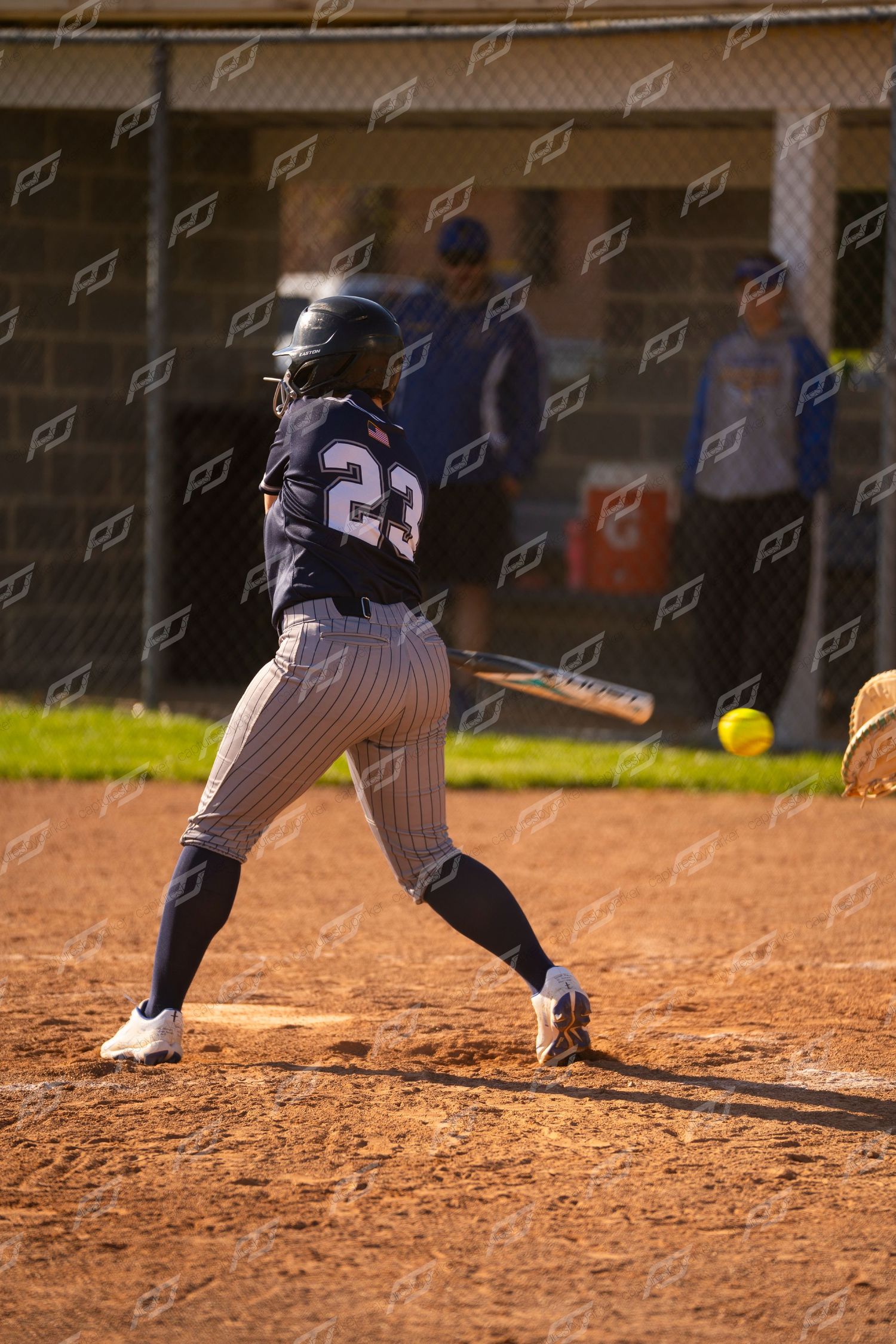 Softball player number 23 swings at a yellow ball during a game on a dirt field.