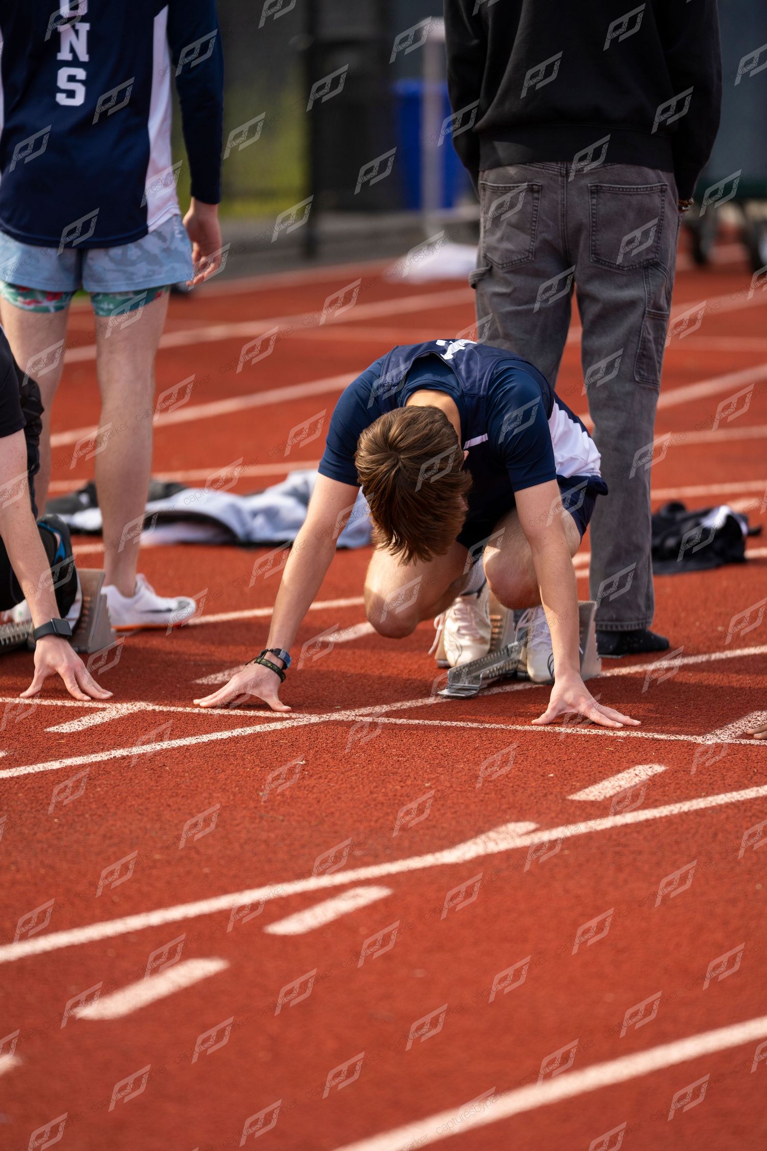 Male athlete crouches in starting position on red track at outdoor track and field meet.