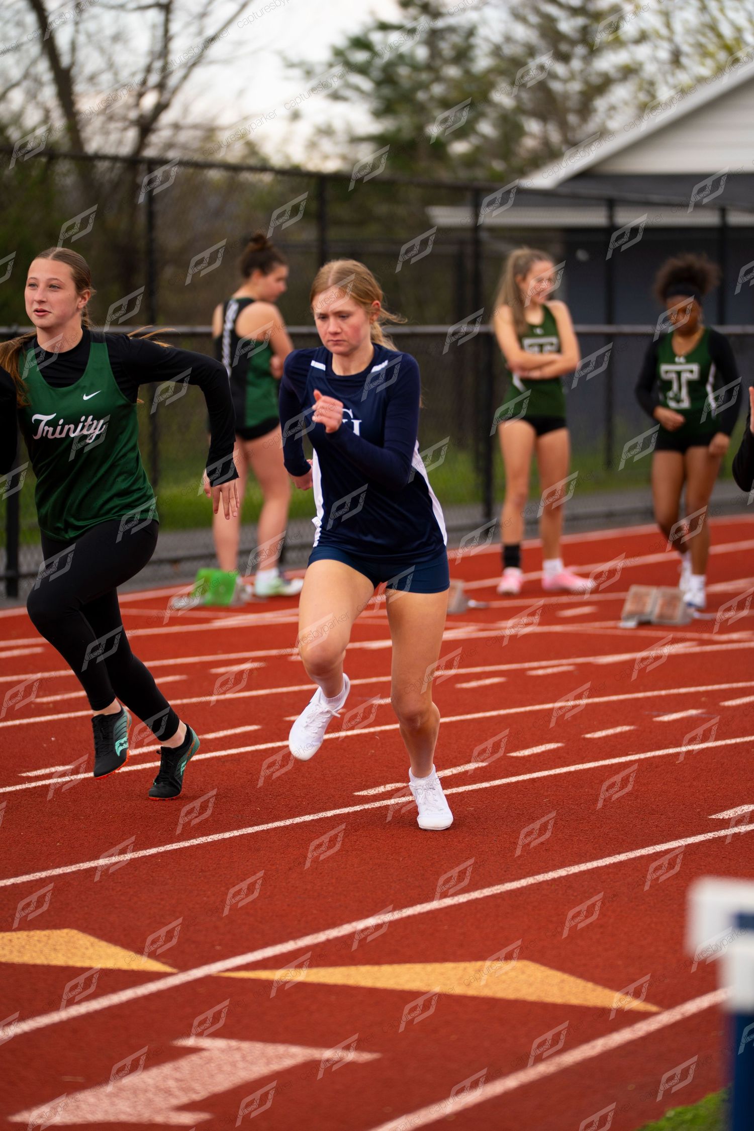 Female sprinter in navy uniform leads race on red track during outdoor high school track meet.