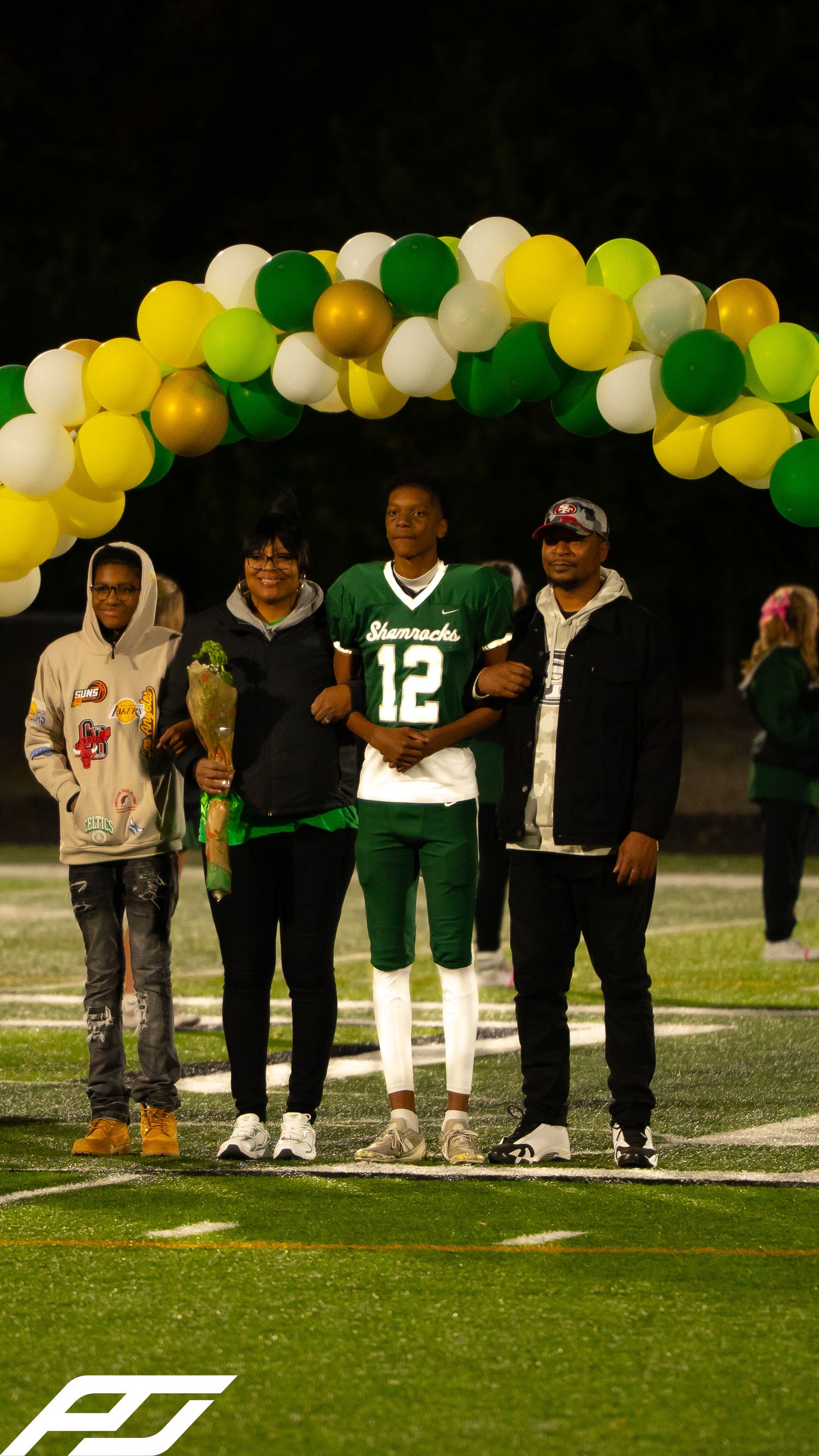 Shamrocks football player number 12 poses with three family members under a balloon arch on field.