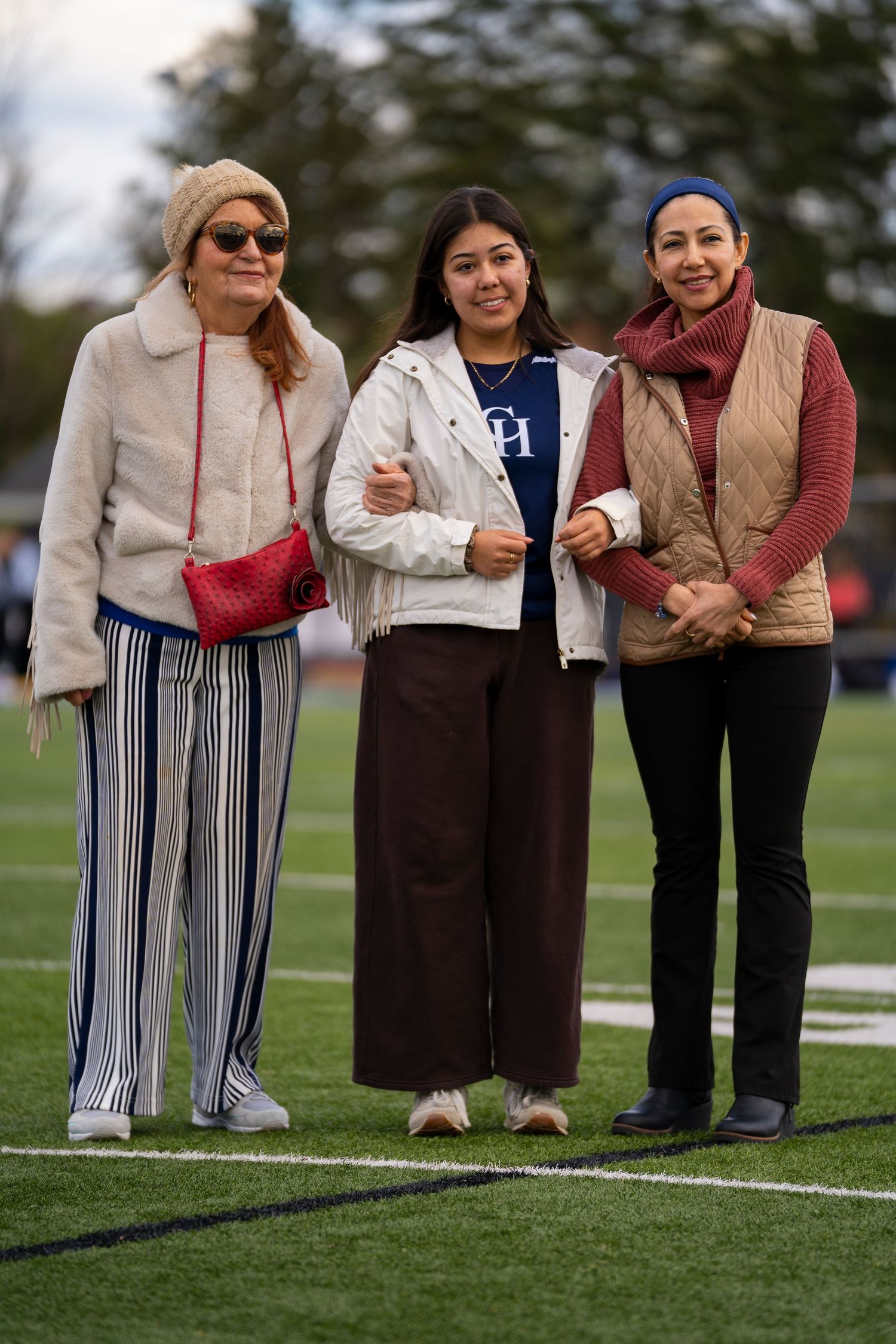 Three women smile together on a football field wearing stylish fall outfits in neutral and warm tones.