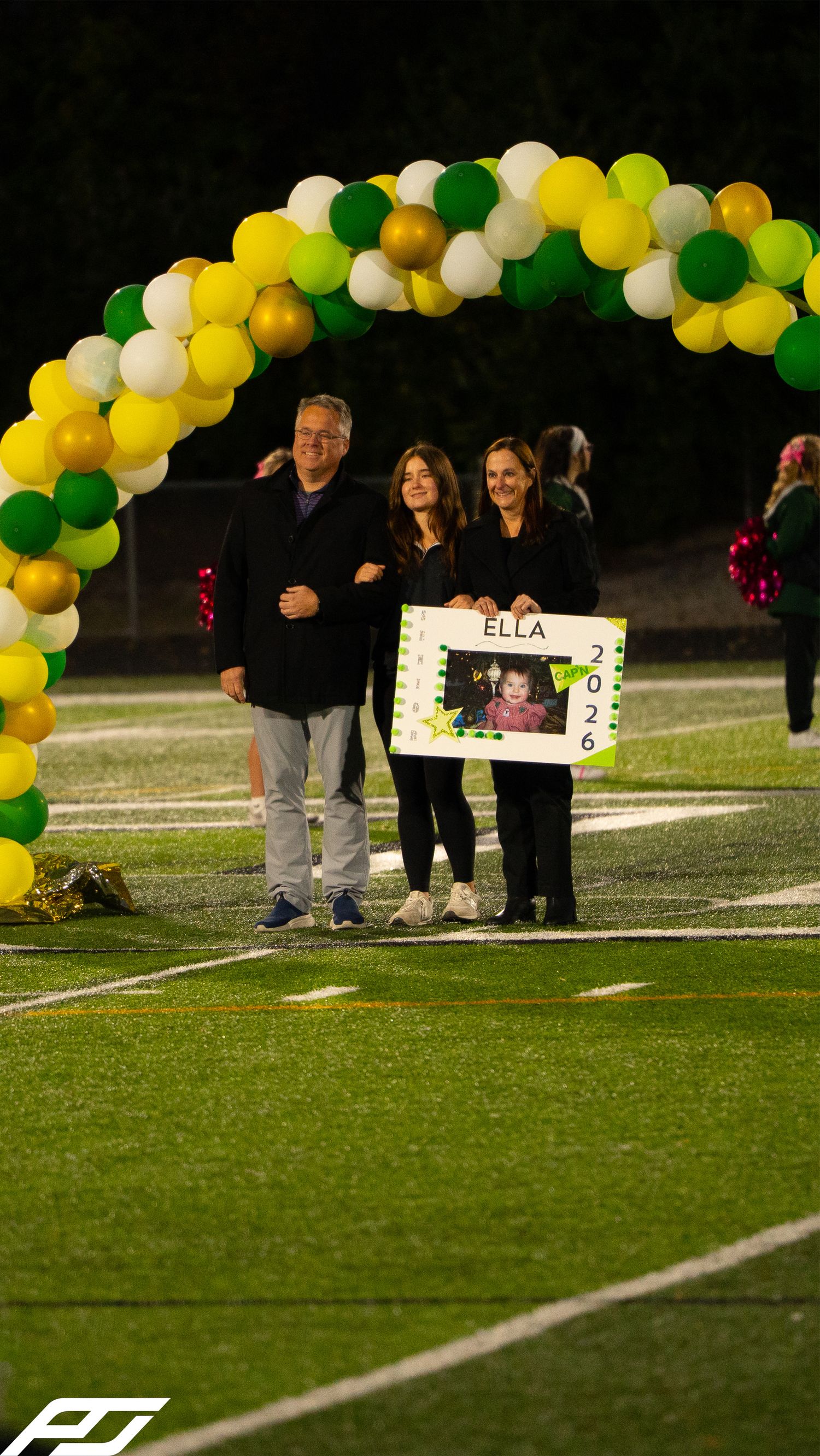 Family holds a senior night poster for Ella 2026 under a green and gold balloon arch on a football field.