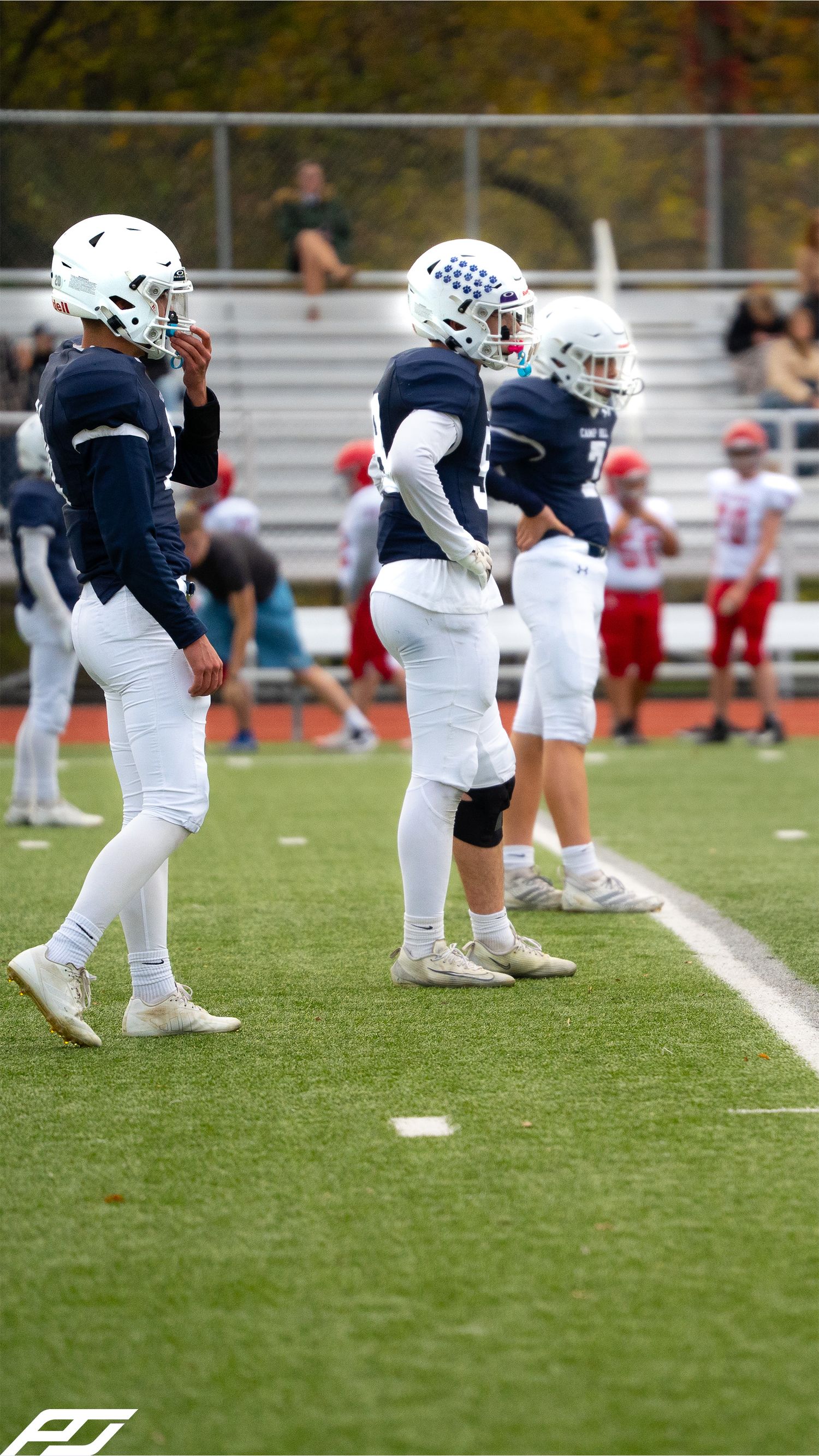 Three youth football players in navy blue uniforms and white helmets stand on the sideline during a game.