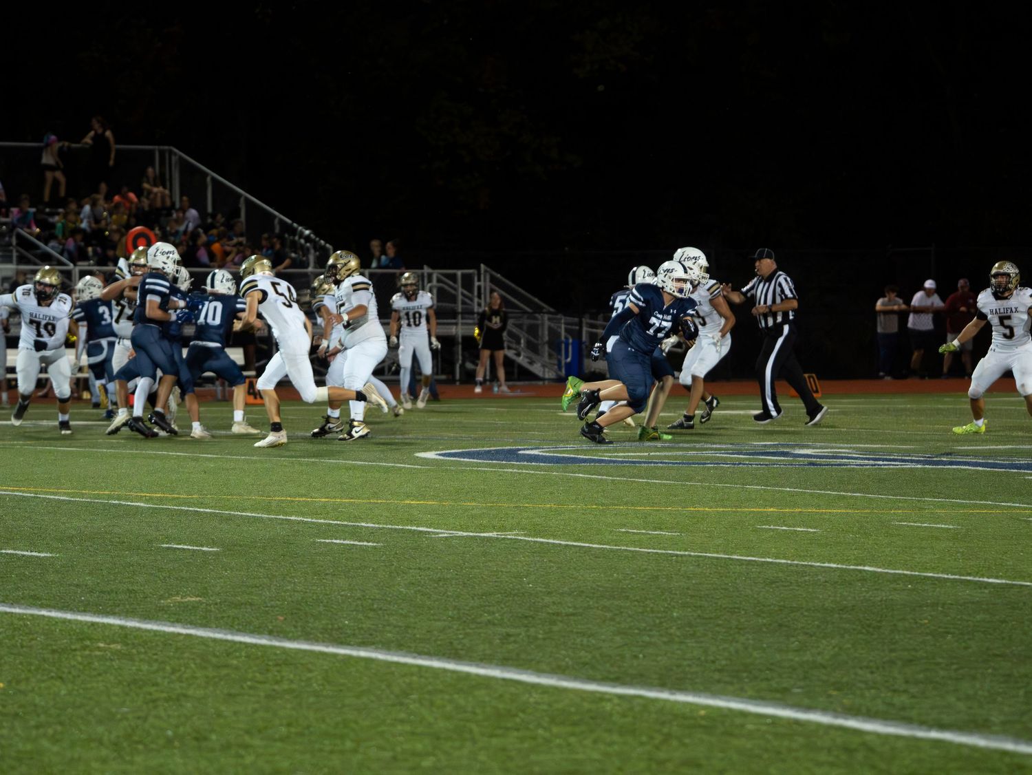 High school football teams clash during a night game, with players in blue and gold uniforms battling at the line.