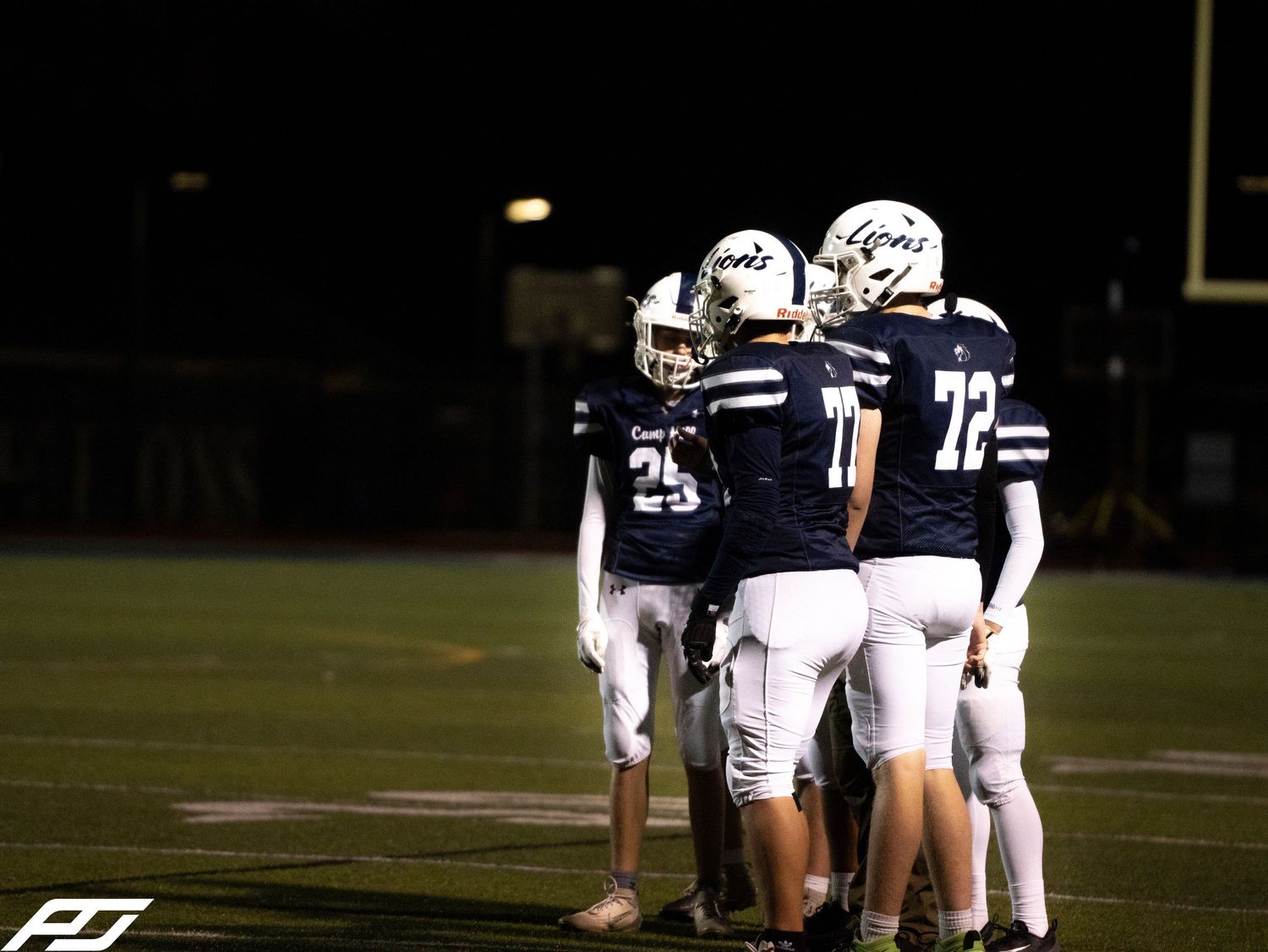 Three Camp Hill Lions football players huddle together on the field during a night game.