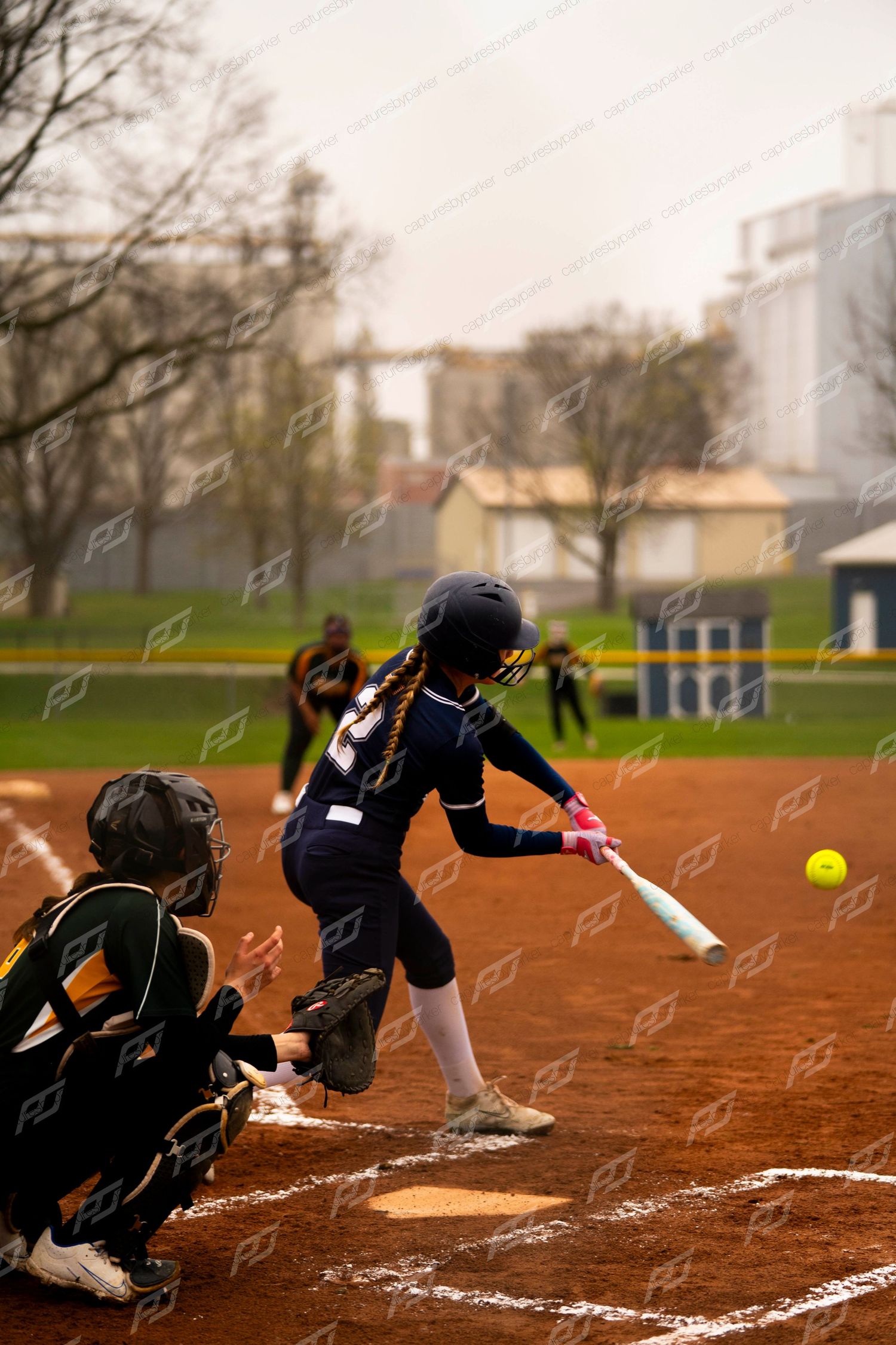 Softball batter in navy uniform makes contact with yellow ball as catcher watches during a game.