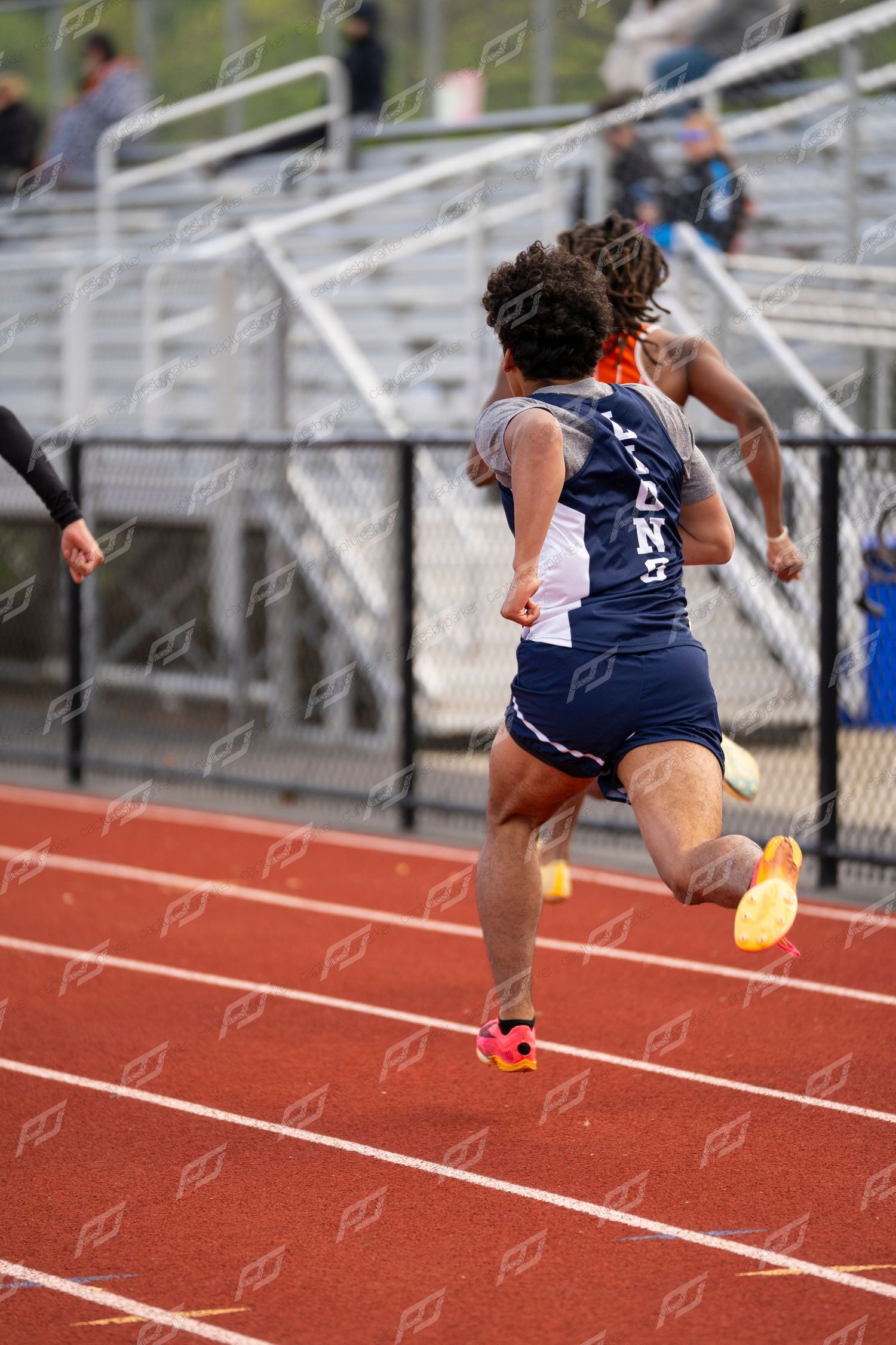 Male track athlete in navy uniform sprints down red track during outdoor meet.