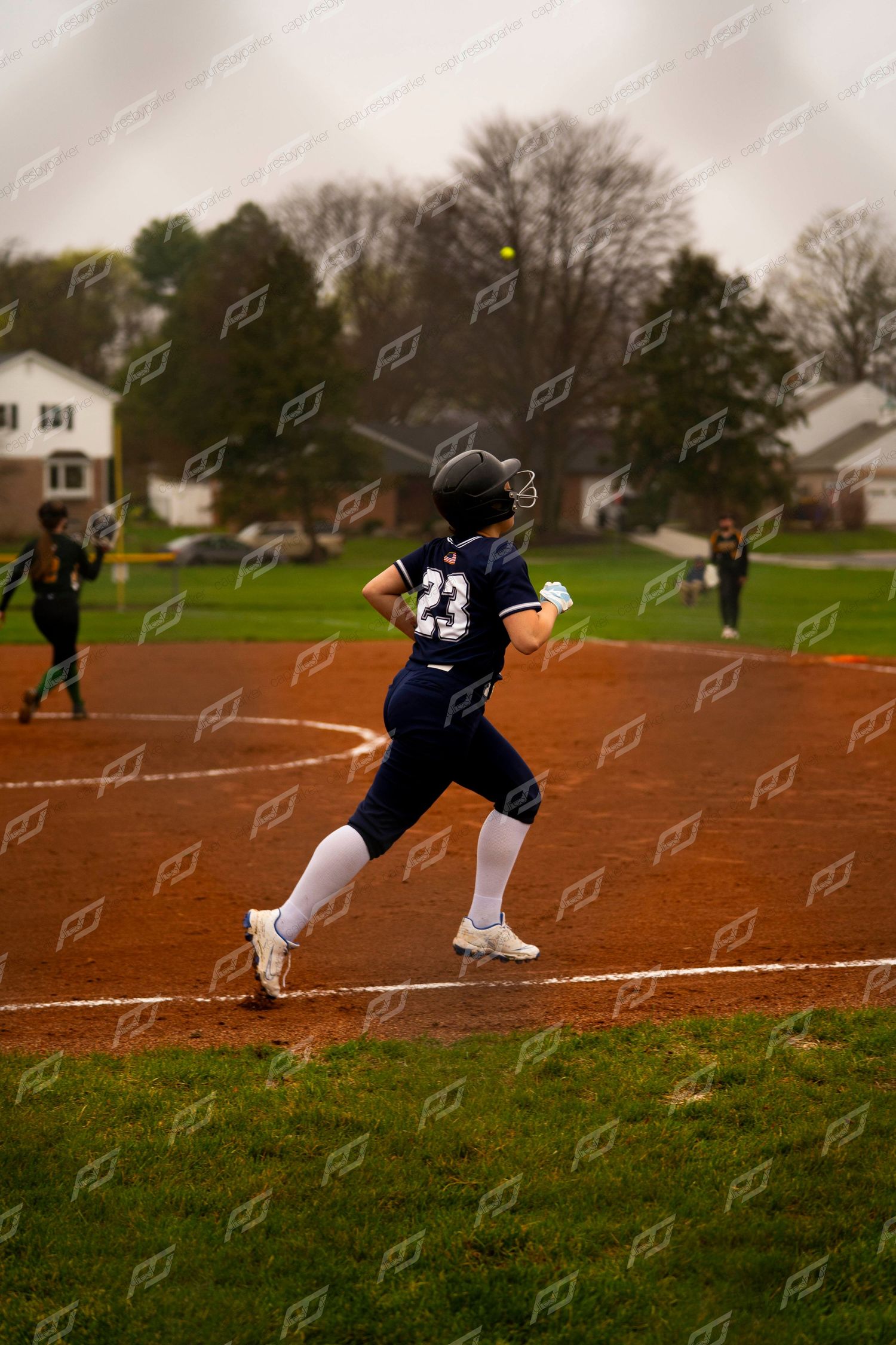 Softball player #23 in navy uniform sprints toward base as ball flies overhead during game.