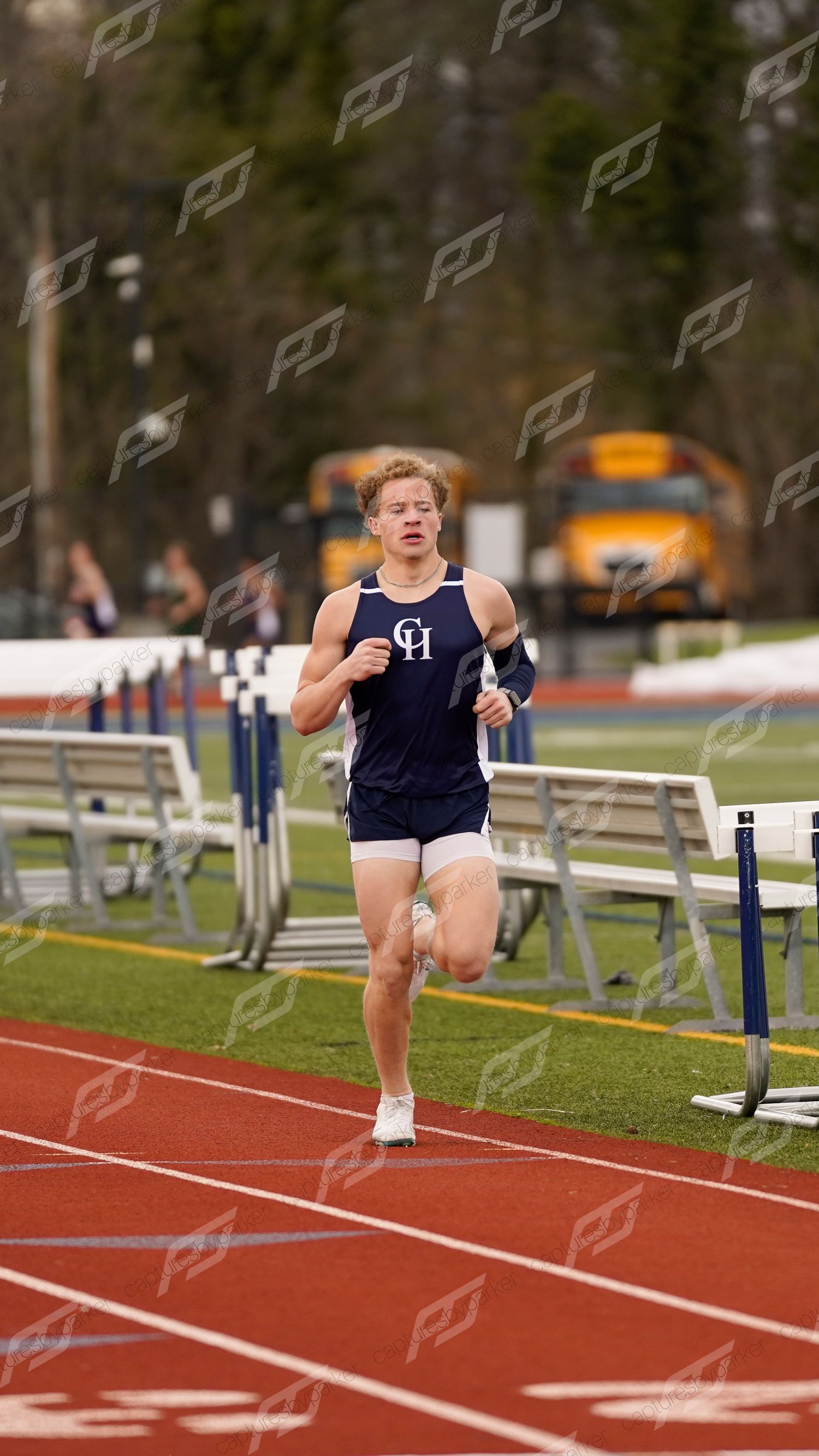 Male high school runner in CH uniform competes on a red track during an outdoor track meet.