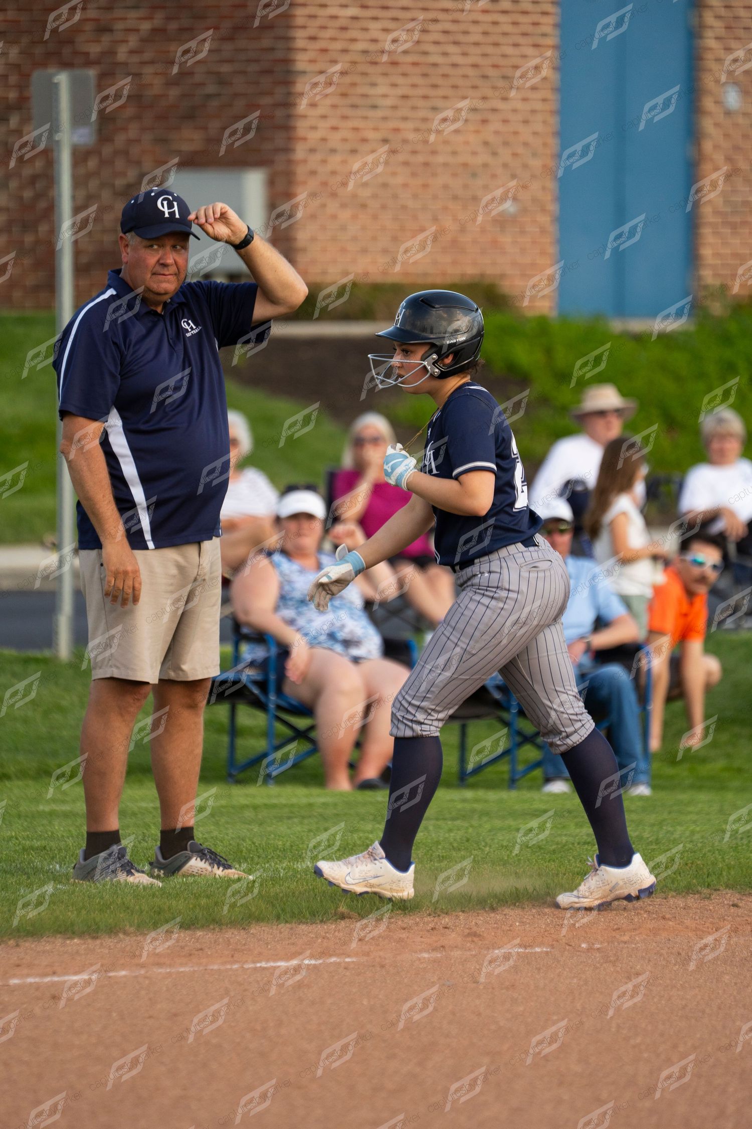 Softball coach signals to female player in navy pinstripe uniform rounding base during game.