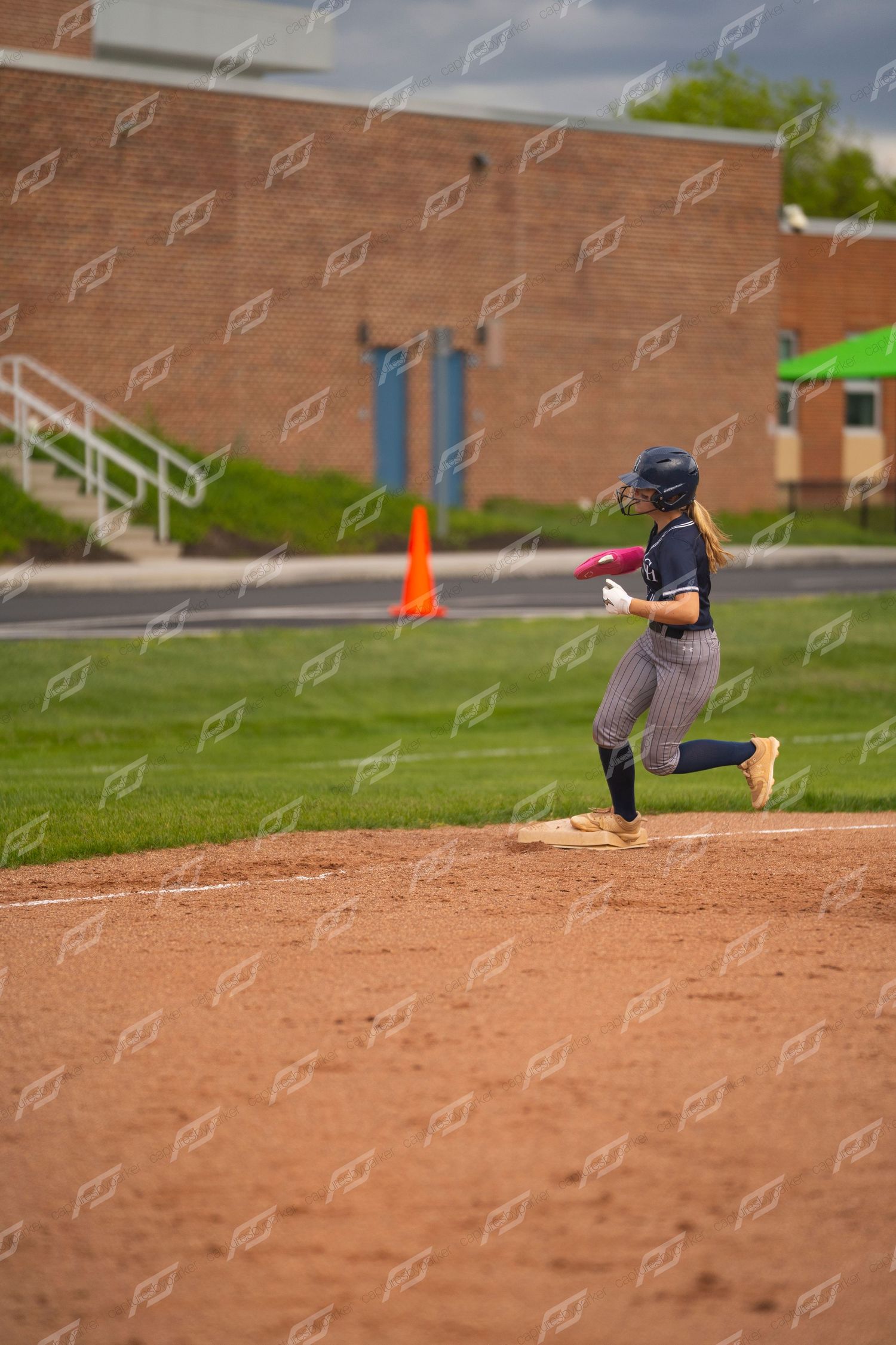 Softball player in navy pinstripe uniform rounds third base during an outdoor game.