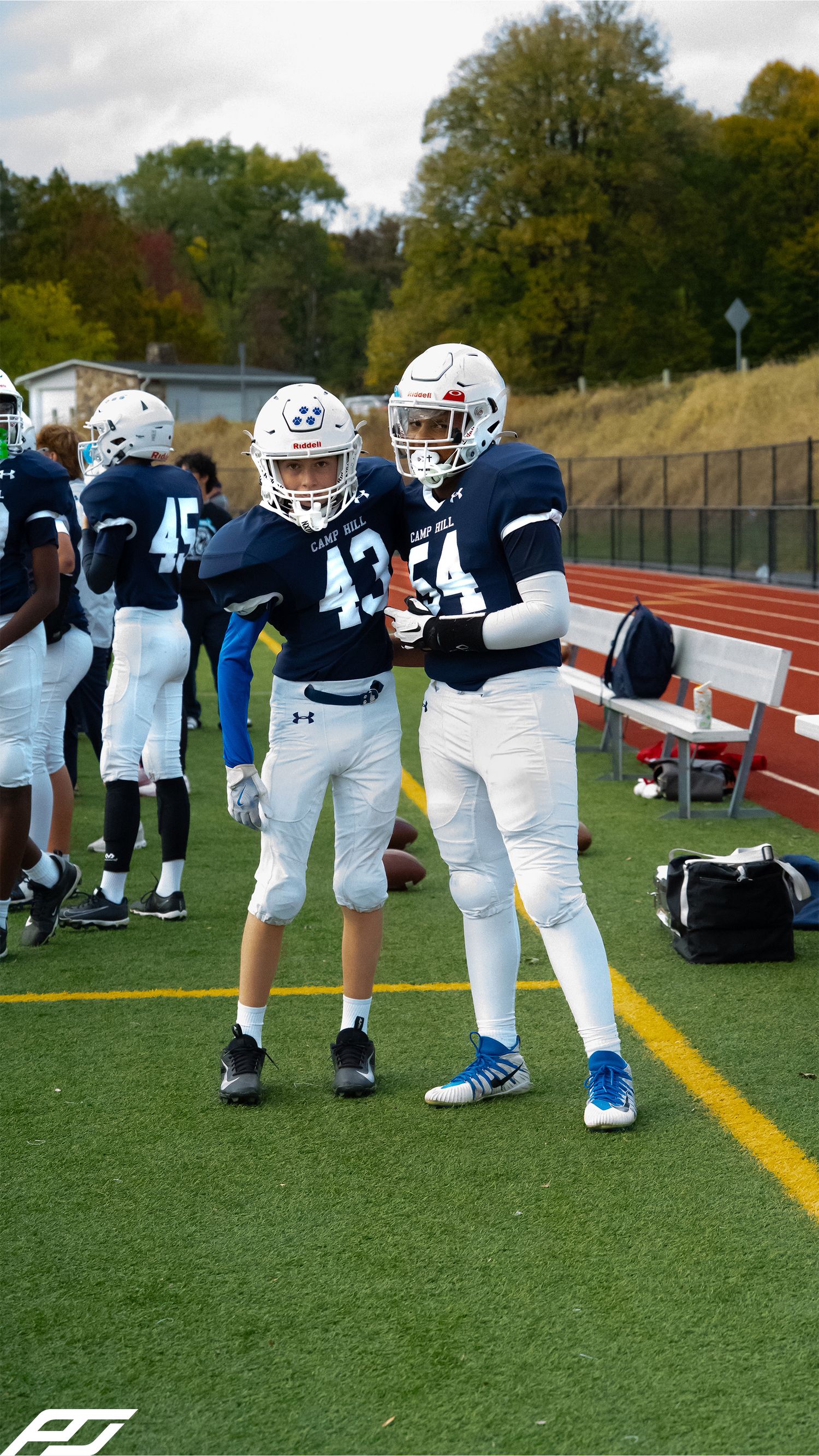 Two youth football players in navy blue uniforms numbered 43 and 54 stand together on a green turf field.