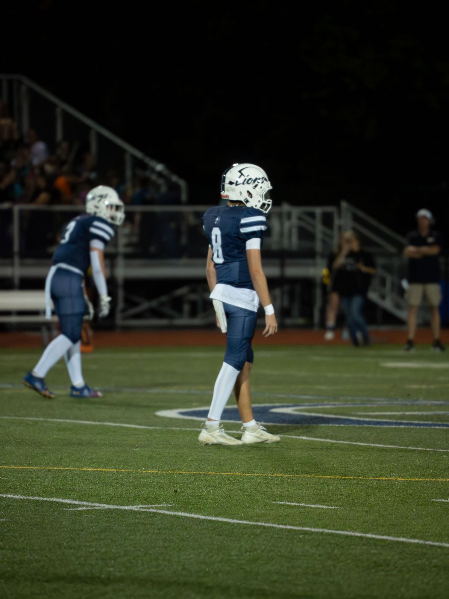 A high school football player wearing number 8 stands on the field during a night game in a white and blue uniform.