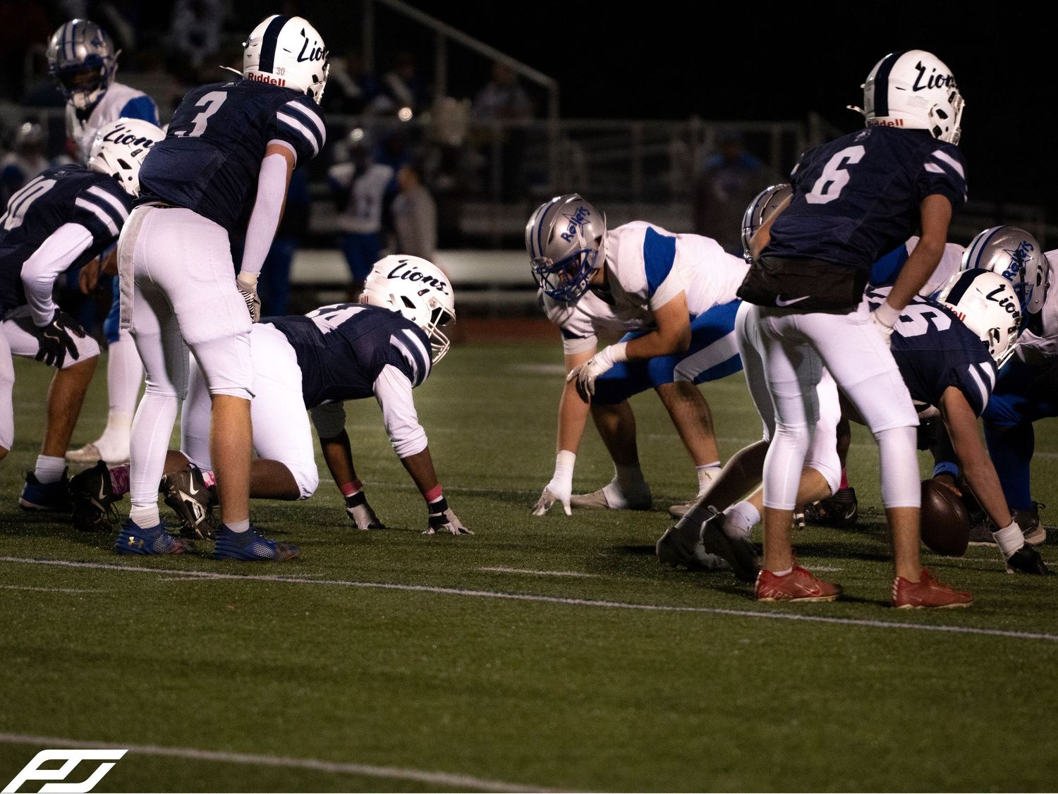 Lions football team lines up against opponents at line of scrimmage during nighttime high school game.