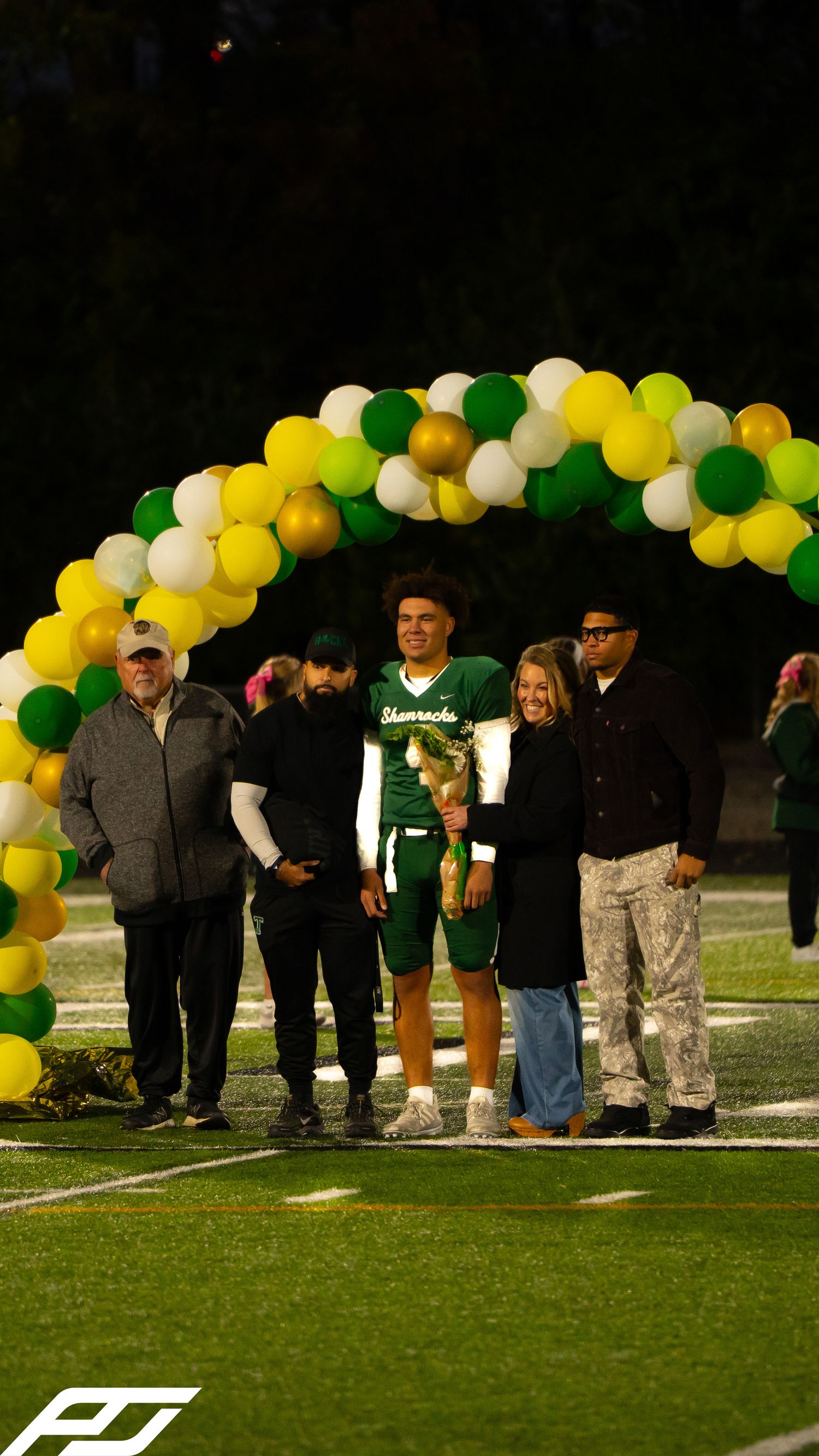 Football player wearing number 1 Shamrocks jersey stands with family under colorful balloon arch at night.