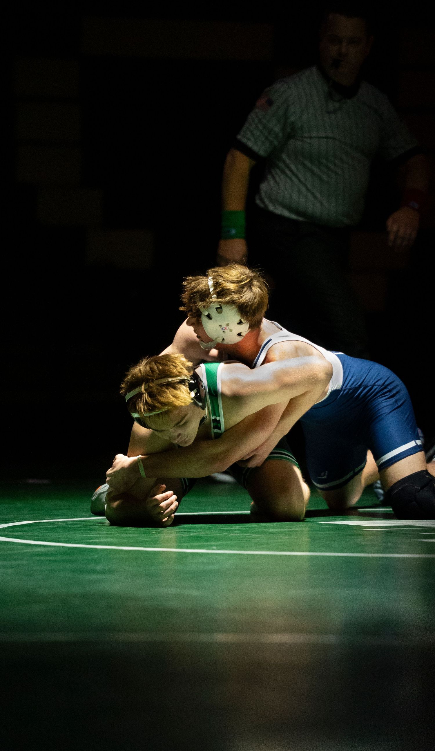 Two wrestlers in blue and green singlets compete on green mat in dramatic lighting.