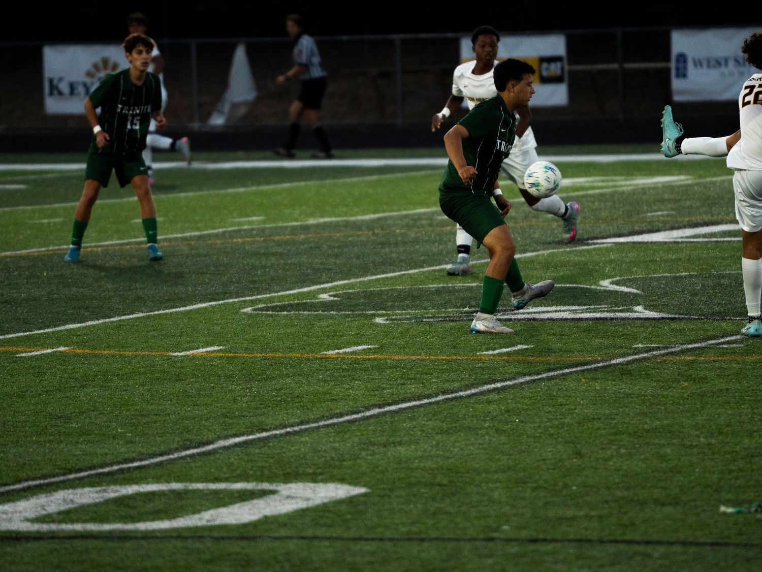 Trinity soccer player controls ball near sideline during evening match under stadium lights on turf field.