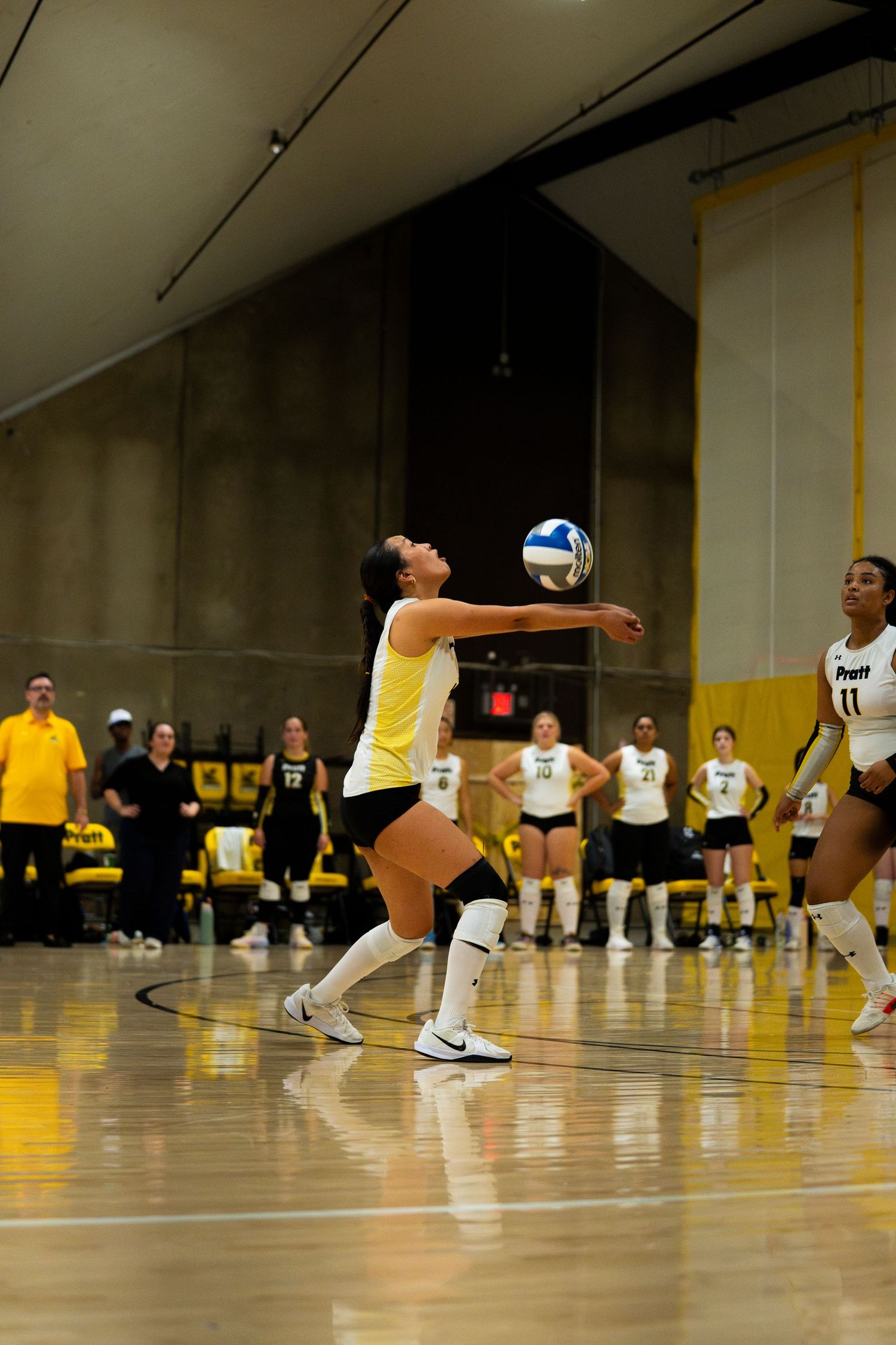 Female volleyball player in yellow uniform performs a dig during an indoor collegiate match with teammates watching nearby.