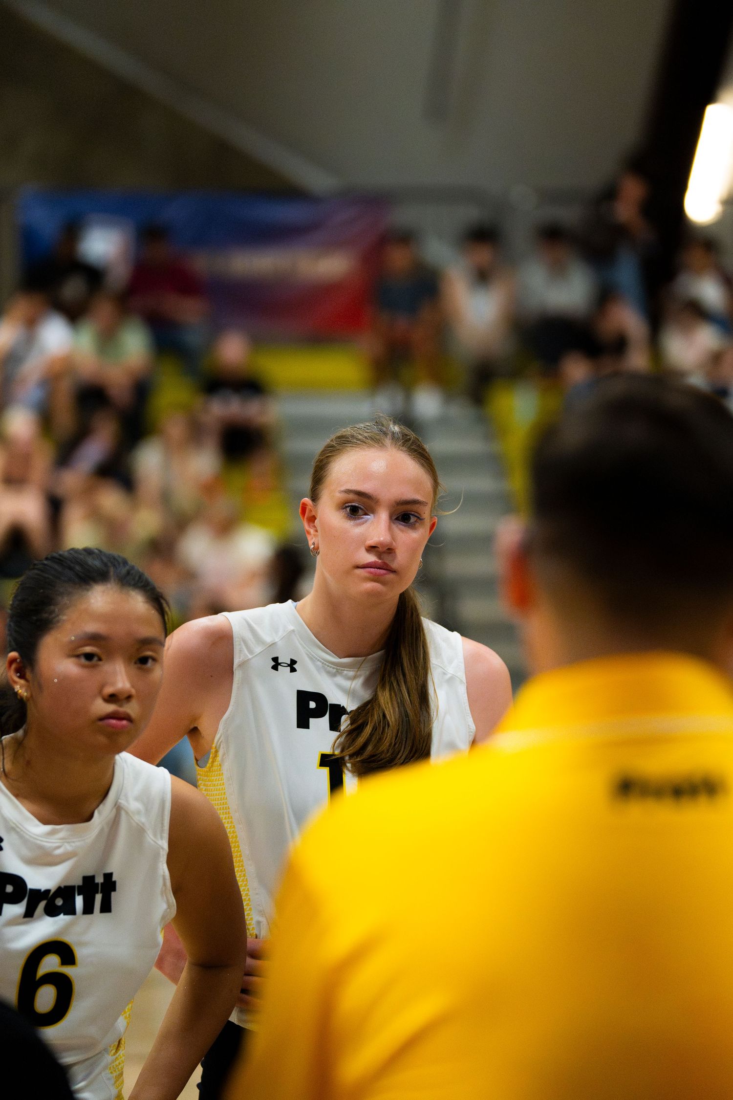 Female basketball players in white Pratt jerseys stand attentively during a game timeout on an indoor court.