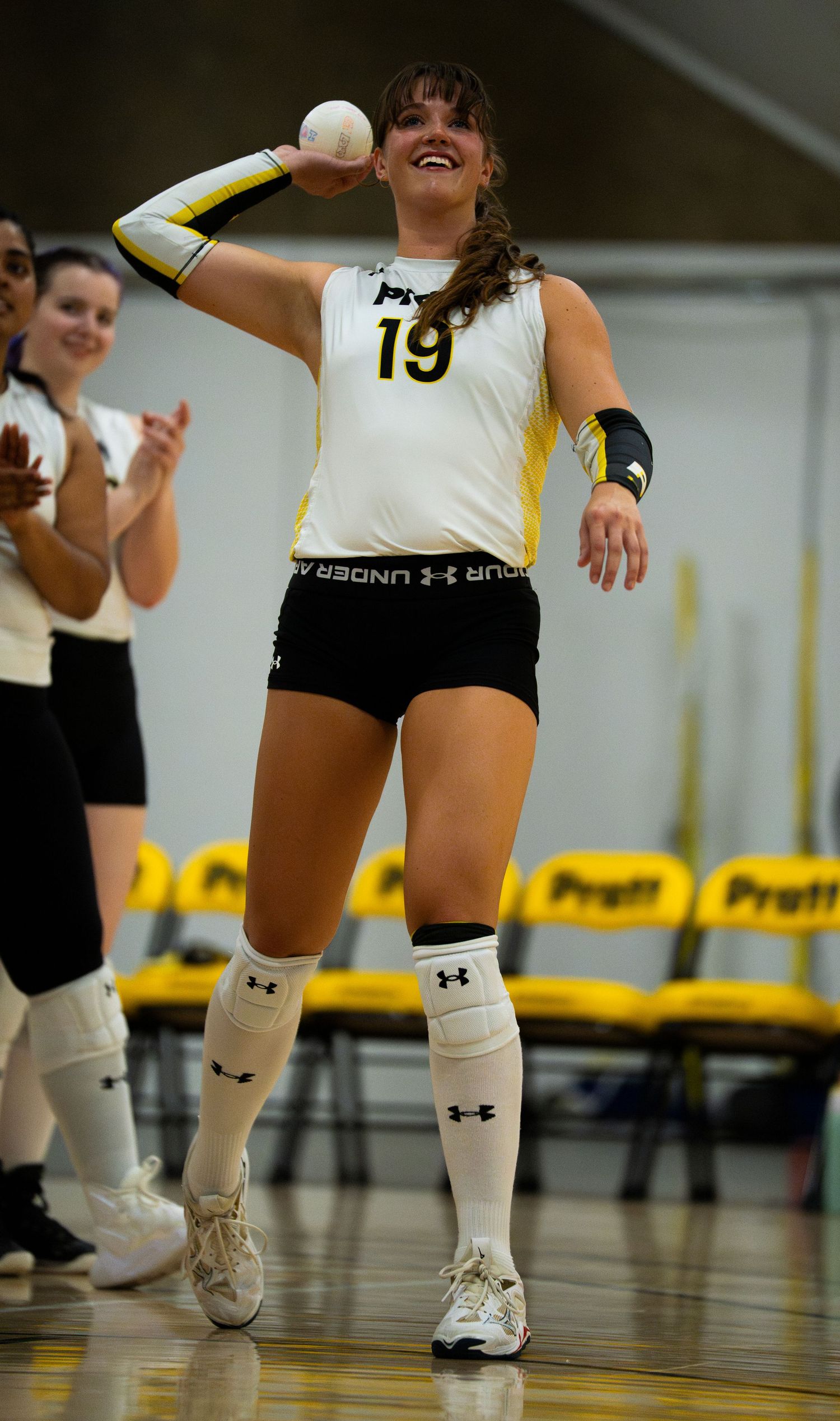 Female volleyball player #19 in white and yellow uniform smiling while holding ball during indoor match.