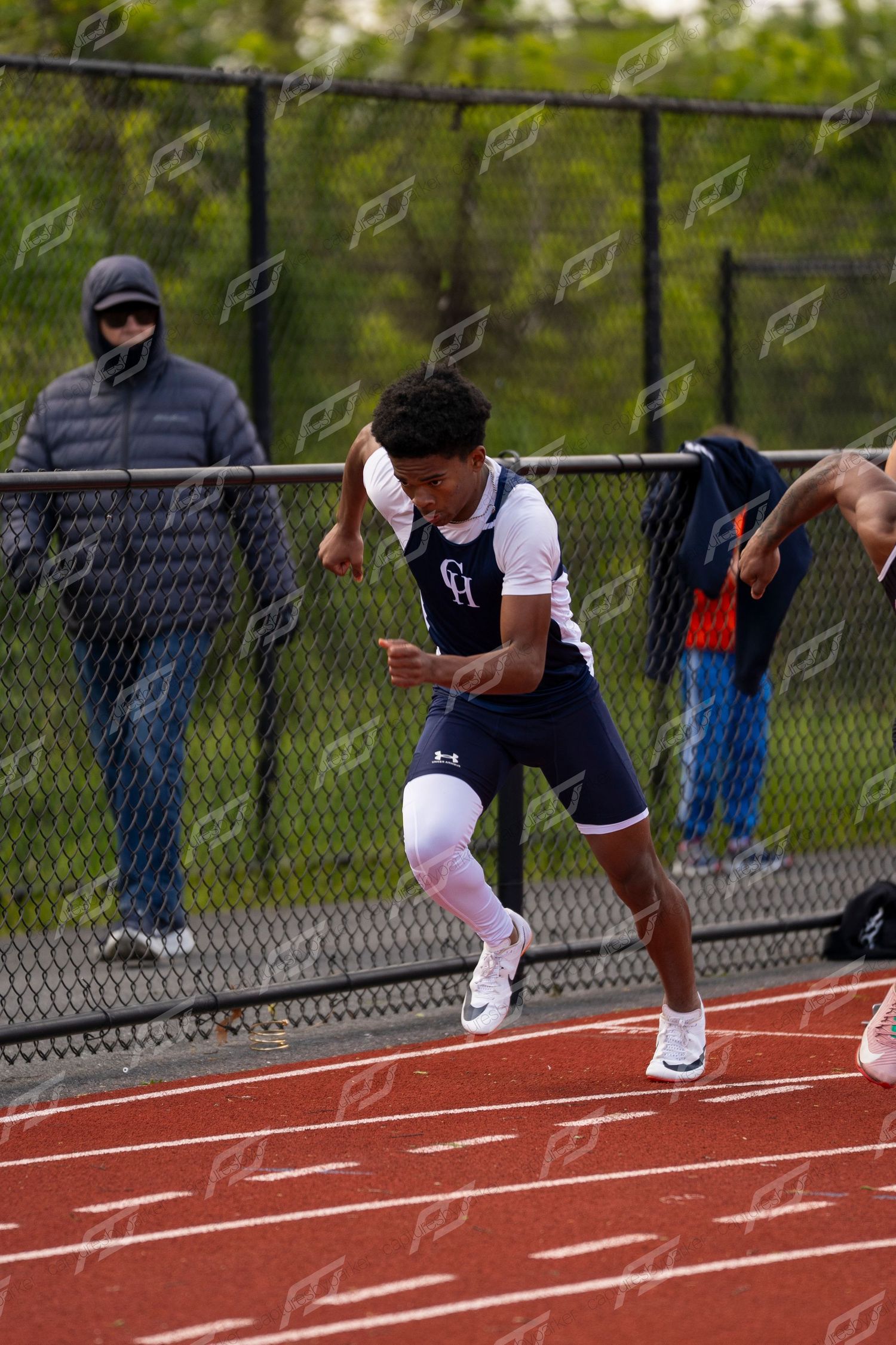 Young male sprinter in navy CH uniform accelerating on red track at outdoor athletic competition.