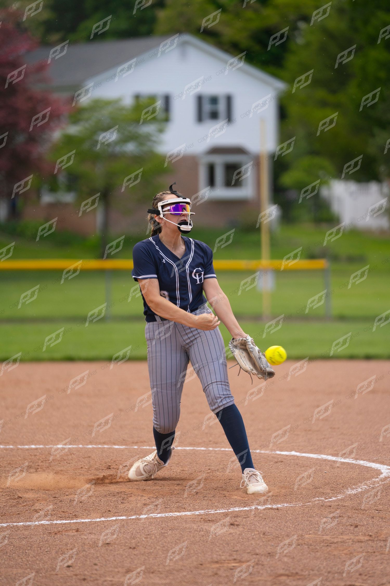 Female softball pitcher in navy pinstripe uniform prepares to release a yellow ball during a game.