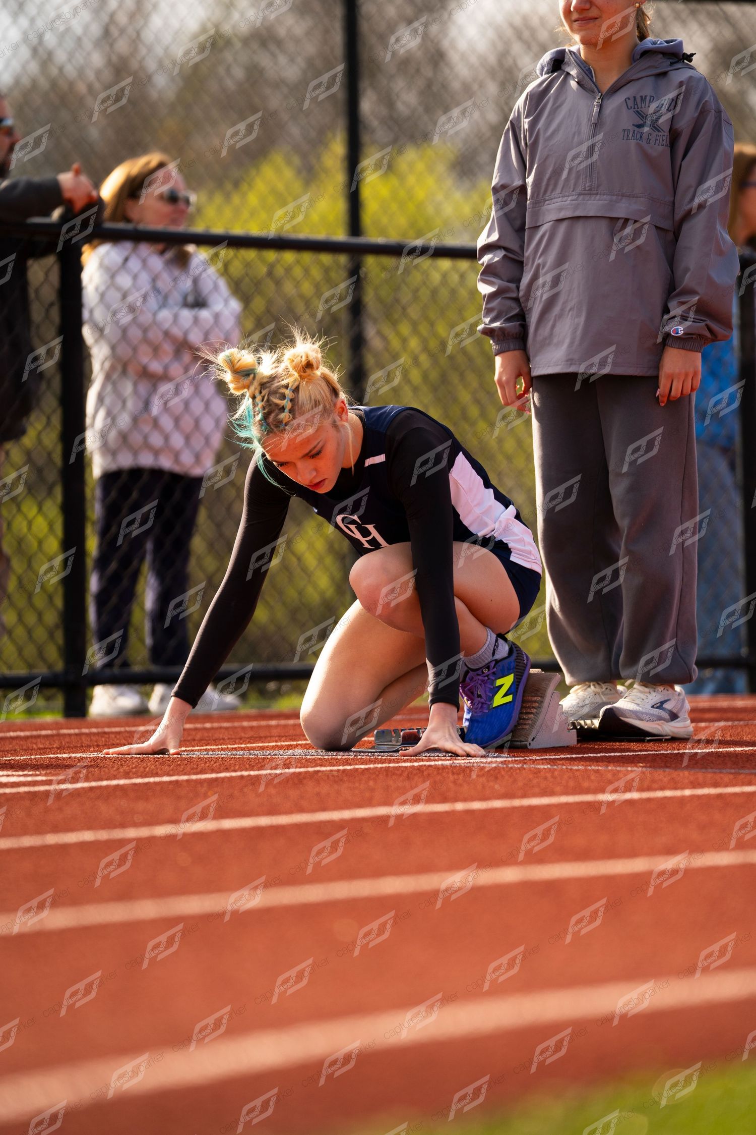 Blonde female sprinter crouches in starting position on red track holding colorful baton at high school meet.