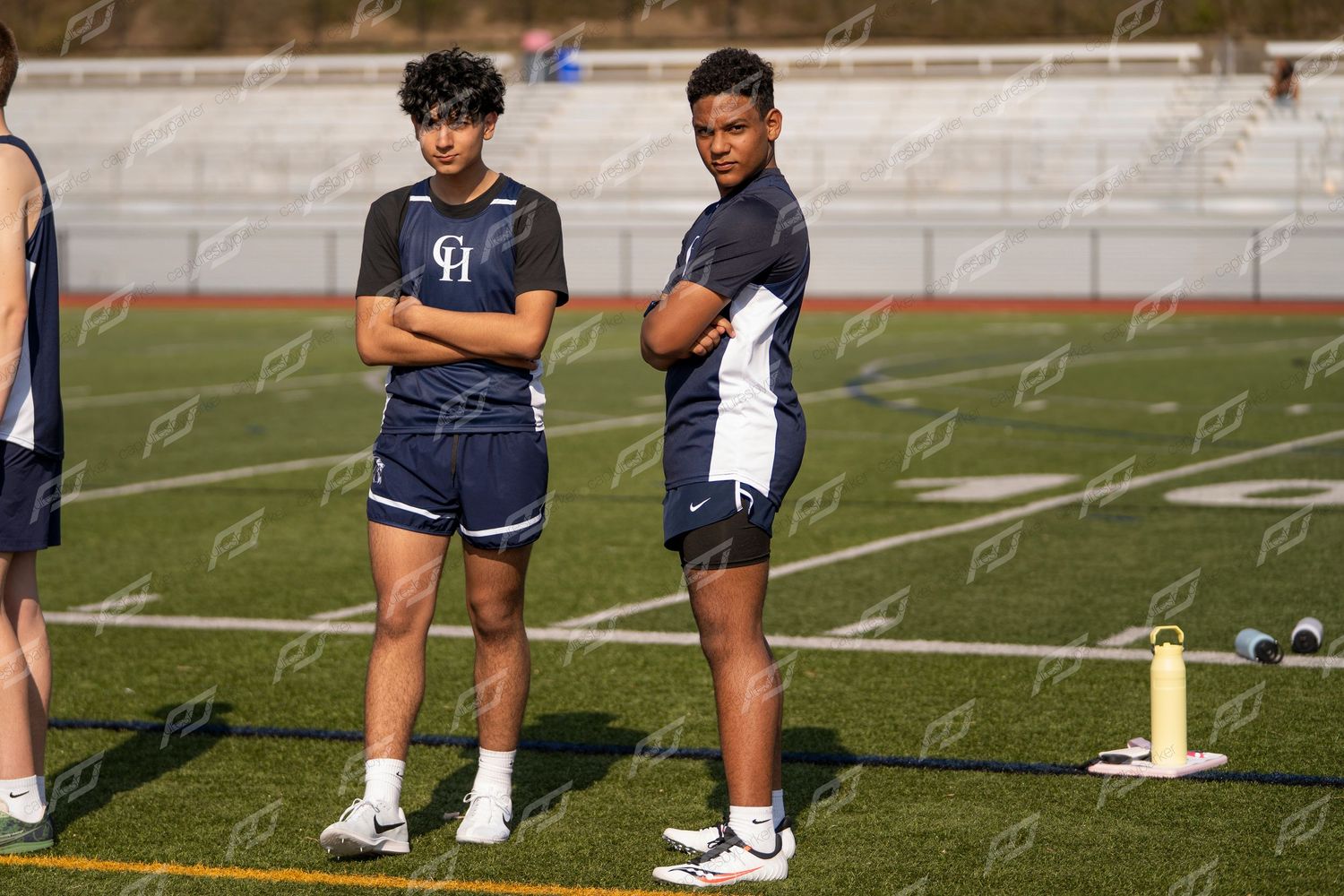 Two male track and field athletes in navy uniforms standing with arms crossed on field.