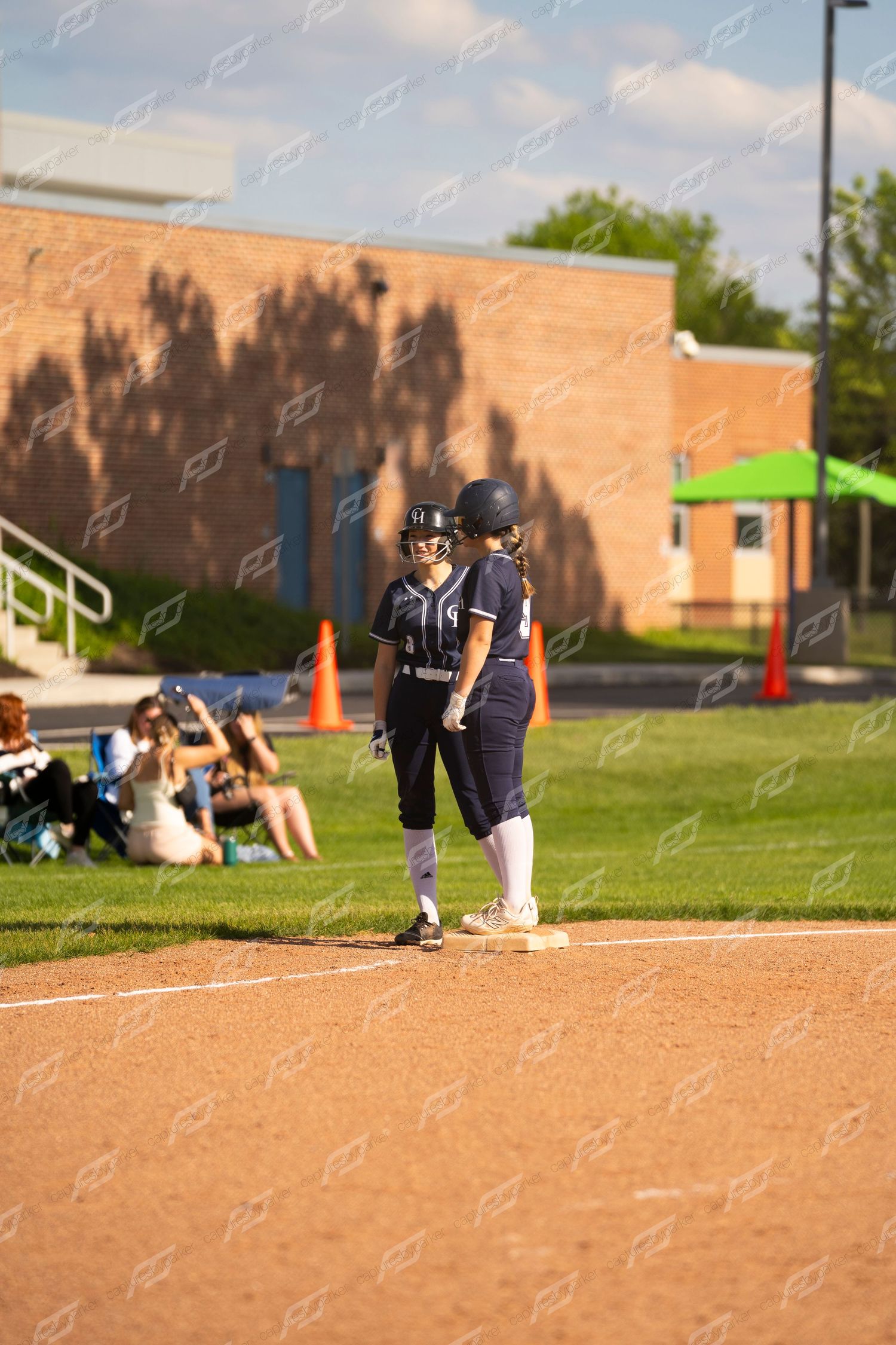 Two softball players in navy uniforms stand at first base during an evening game with spectators in the background.