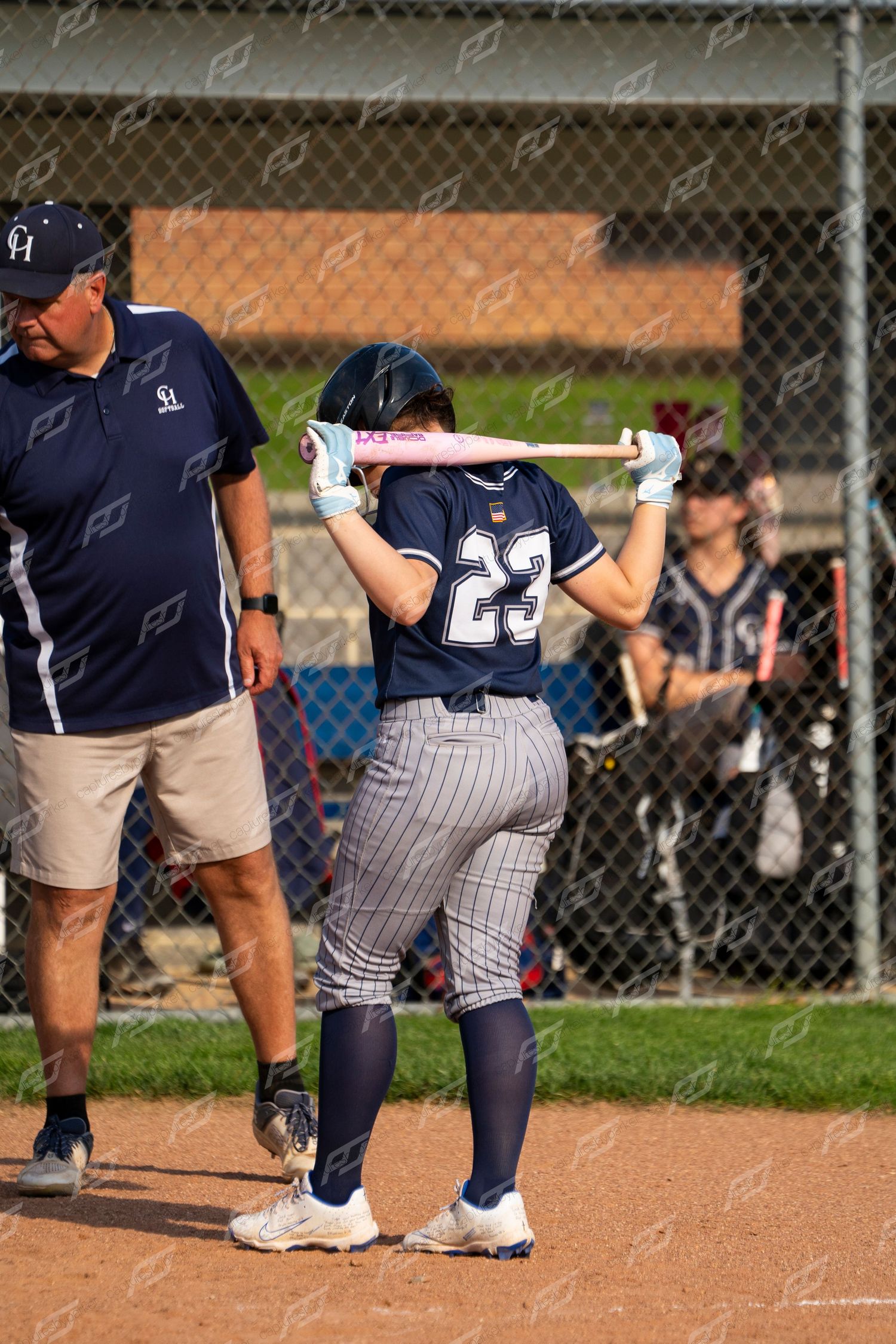 Softball player number 23 in navy uniform rests bat on shoulders near coach during game.