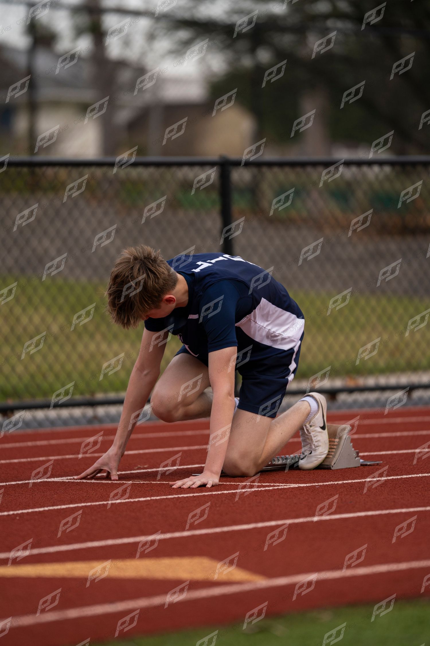 Male sprinter in navy uniform crouches in starting position on red track.