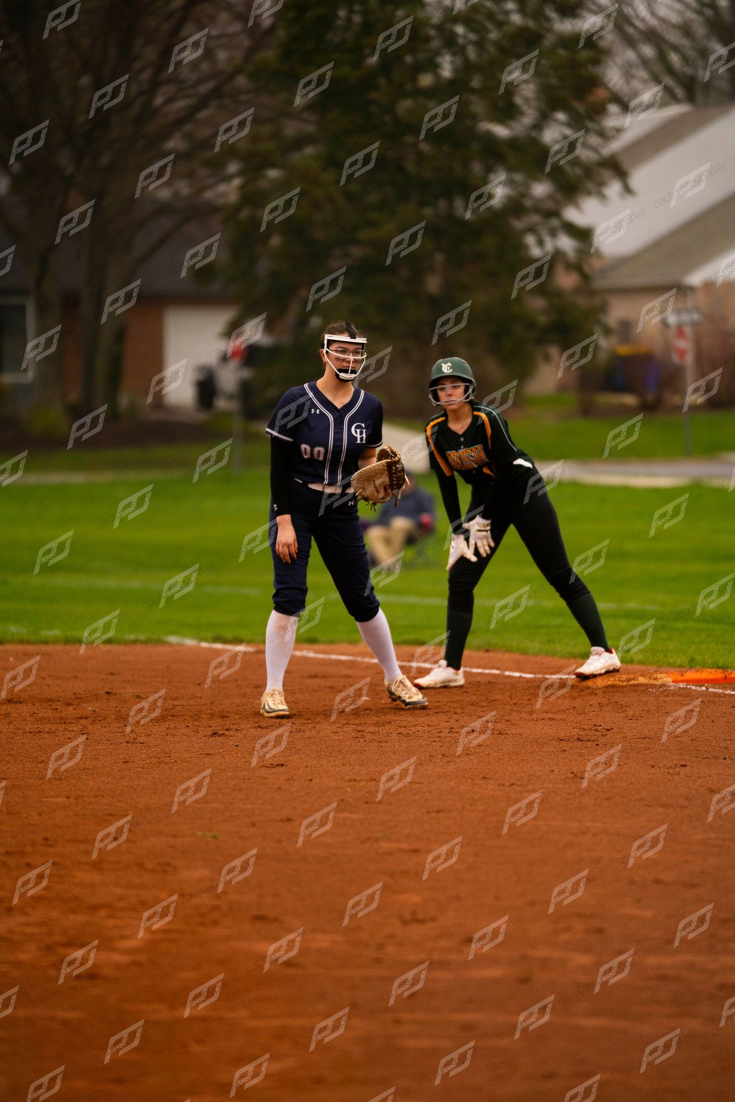 Navy uniform fielder and opponent in black uniform stand ready at base during softball game.