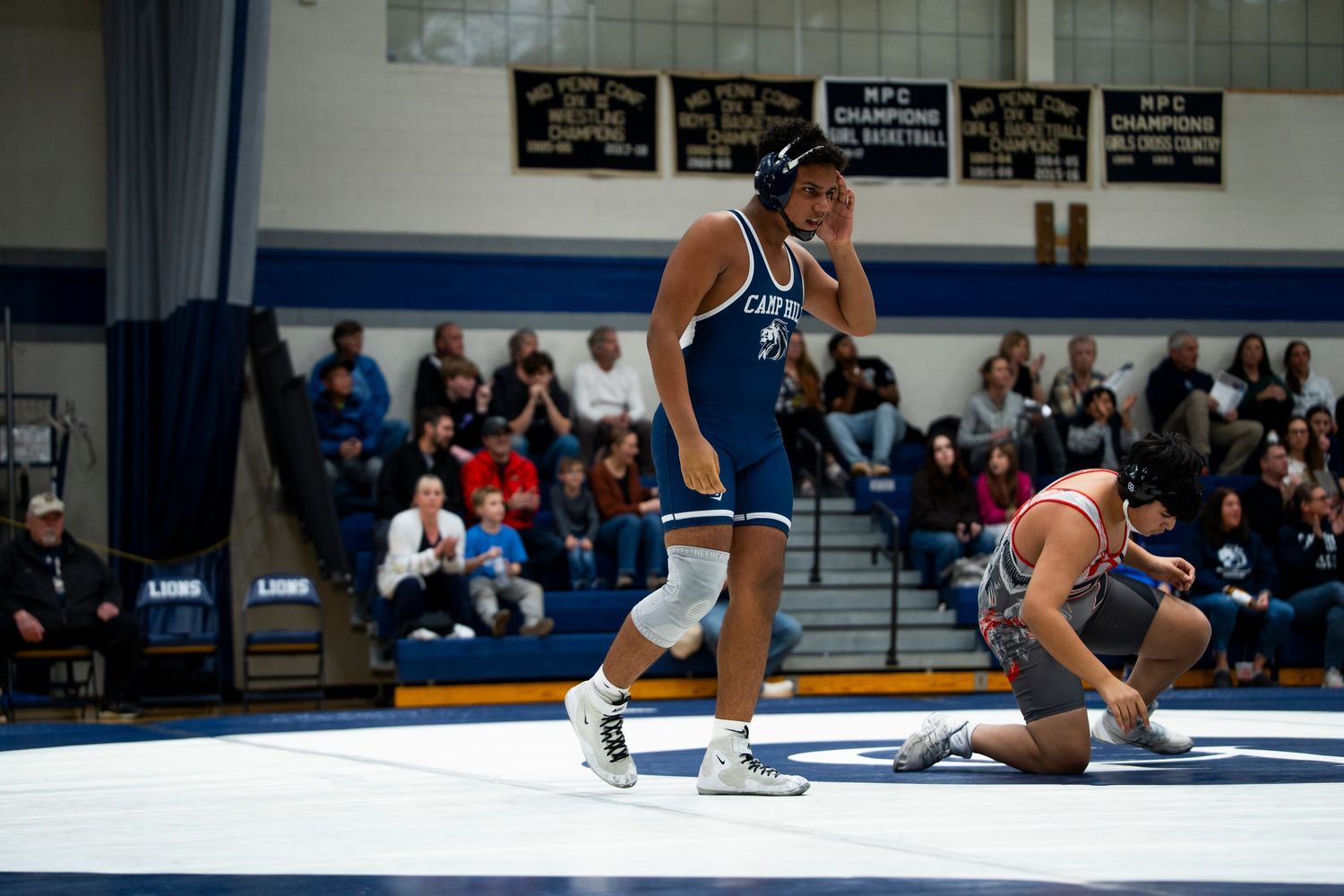 Victorious high school wrestler in blue singlet celebrates win as opponent kneels on wrestling mat.